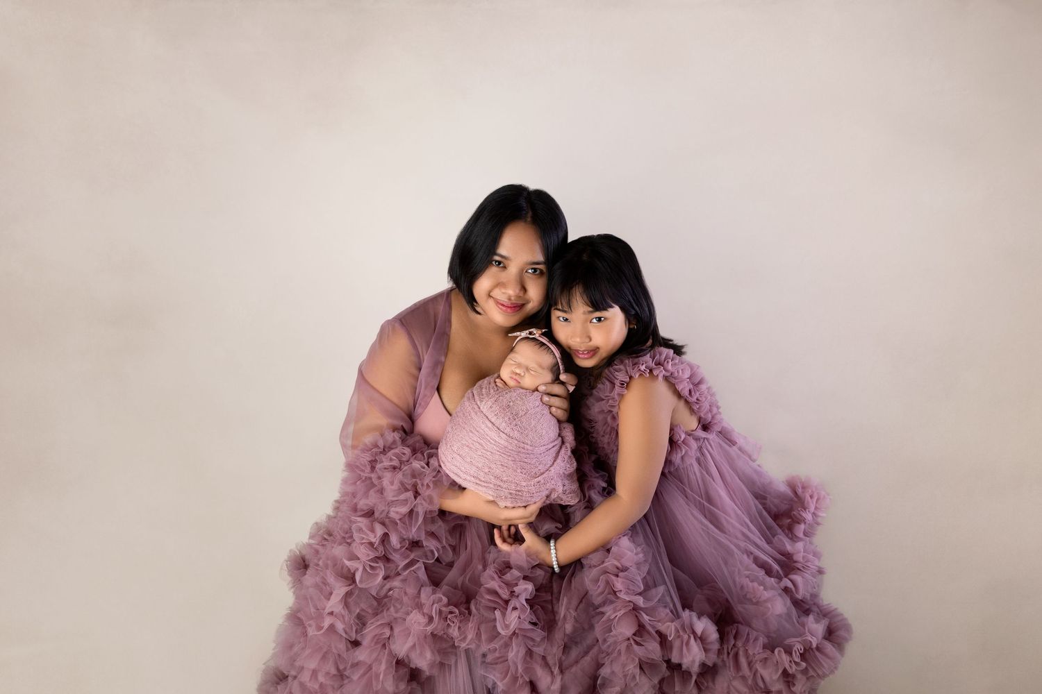 Mother in purple ruffle gown holds newborn while older daughter in matching dress poses for studio portrait.