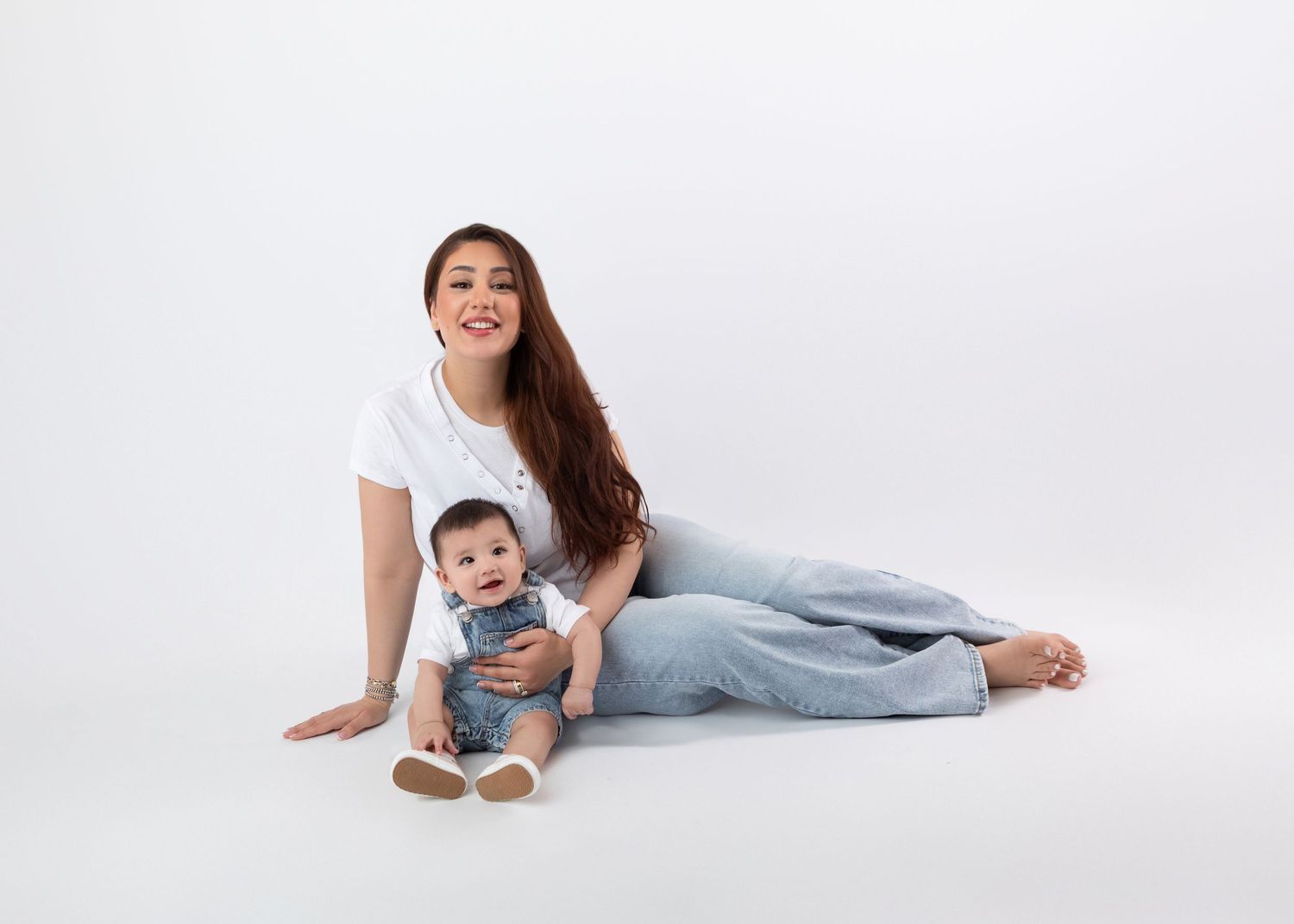 Cheerful mom with long hair sits beside her cute baby boy in a white studio setting.