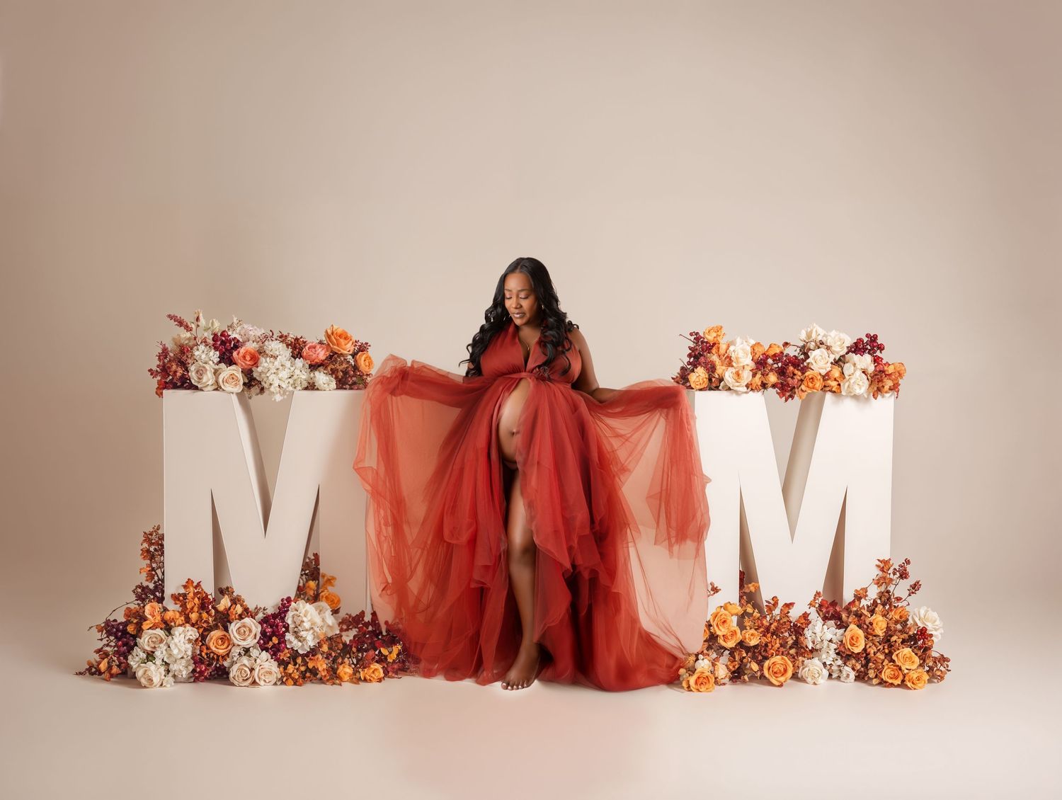 Maternity photo of woman in red tulle dress standing between large MOM letters and flowers.