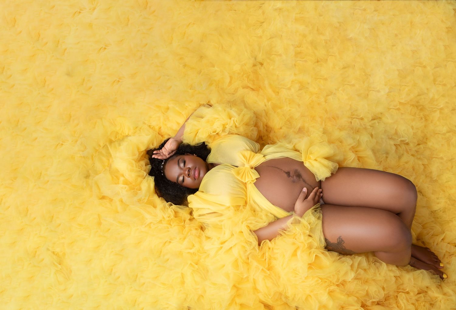 Pregnant woman lying in a sea of yellow tulle fabric during a creative maternity photo shoot.