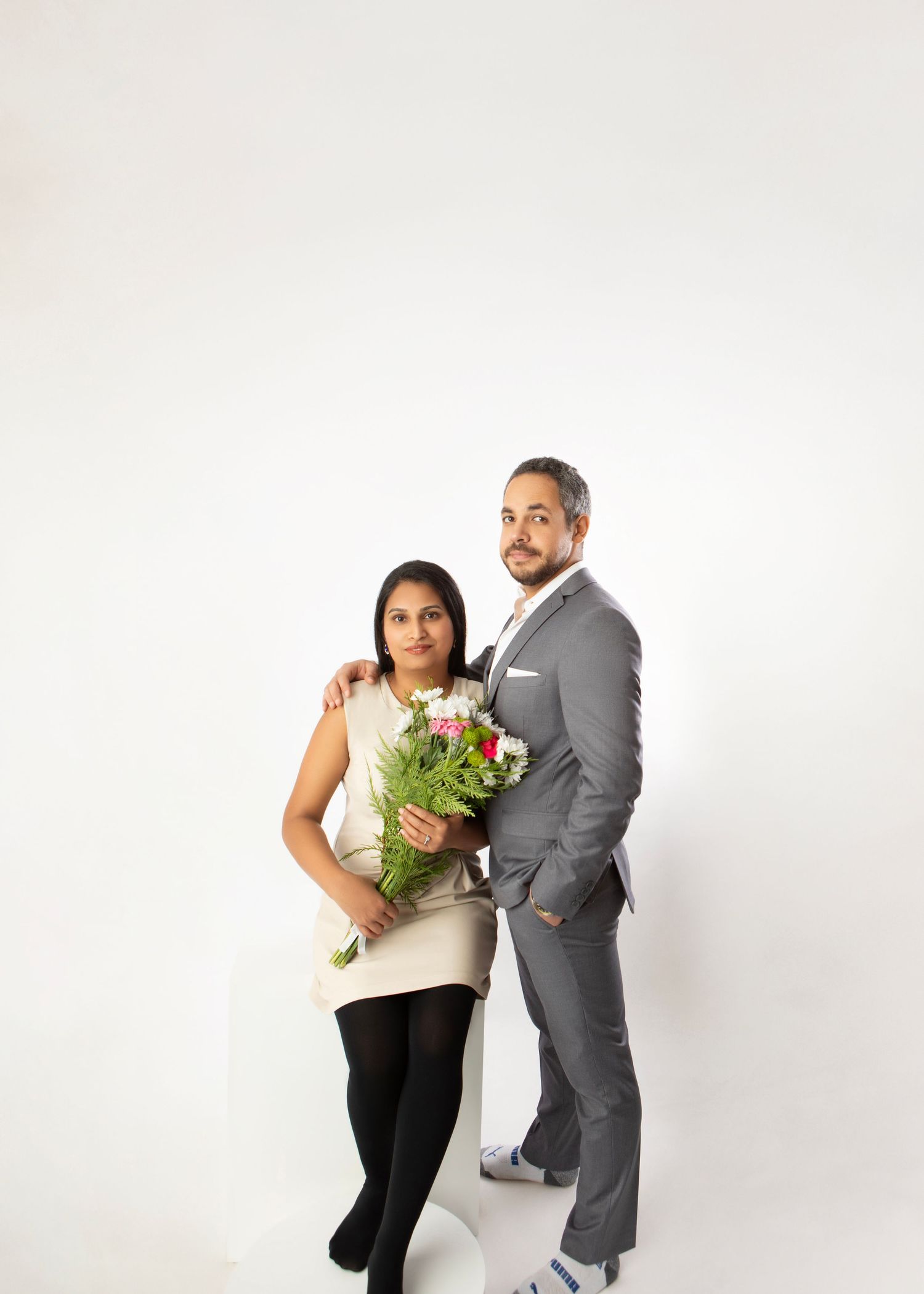 Couple posing in studio, woman holding red and white flowers, man in gray suit with hand on her shoulder.