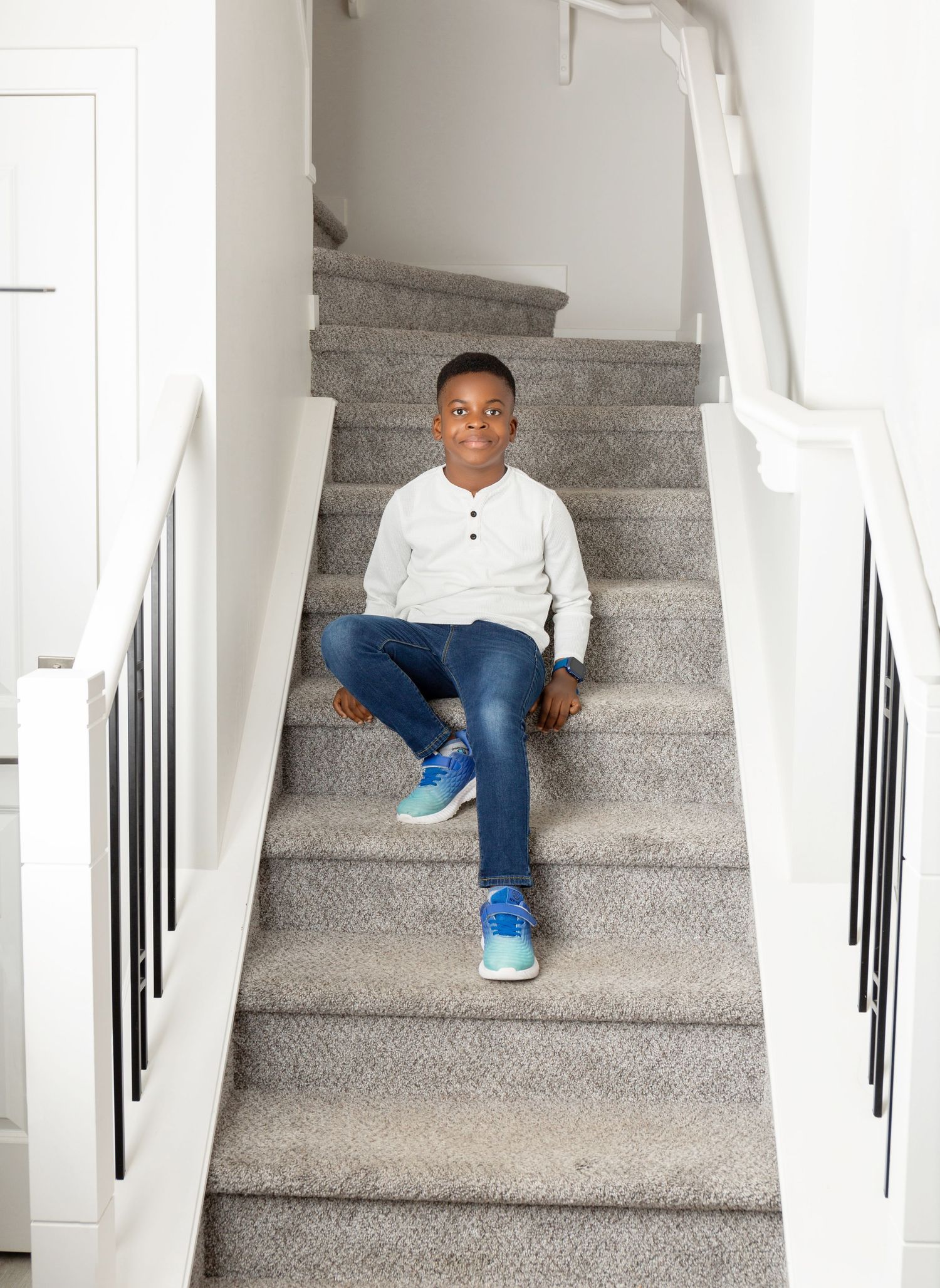 Young boy in white shirt, jeans, and blue sneakers sitting on carpeted stairs with white railings.