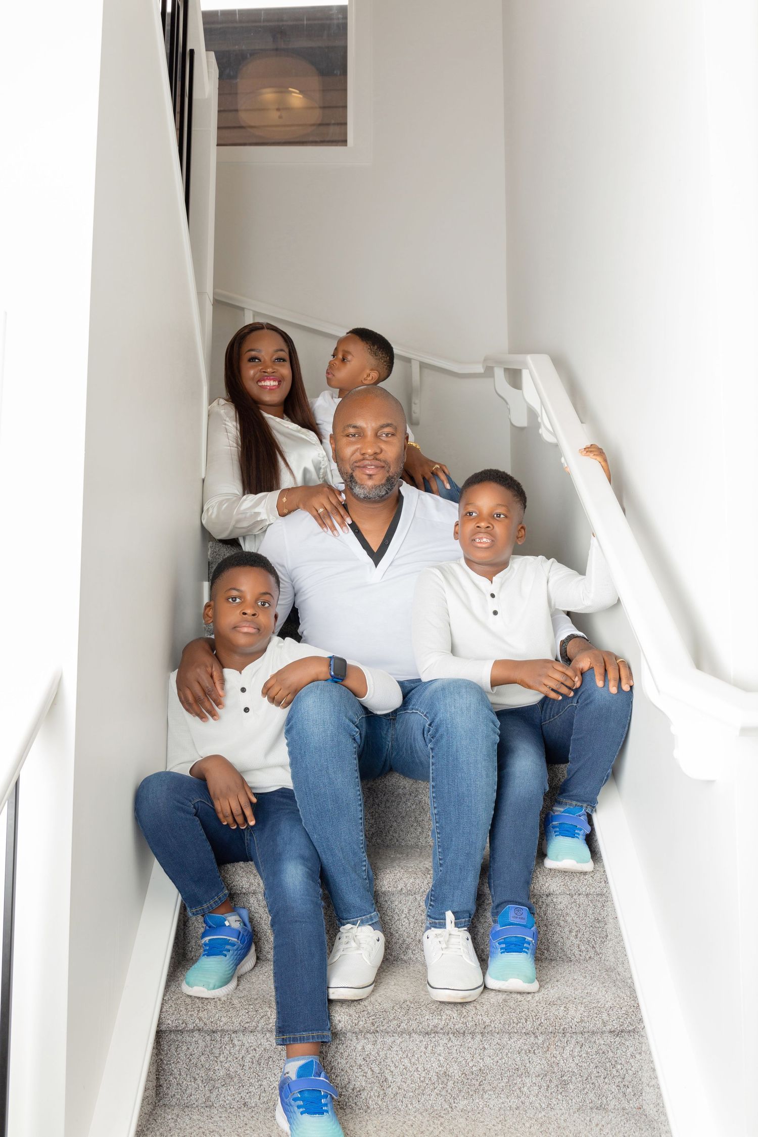 Happy African American family of five posing together on white indoor staircase, wearing matching white tops and jeans.