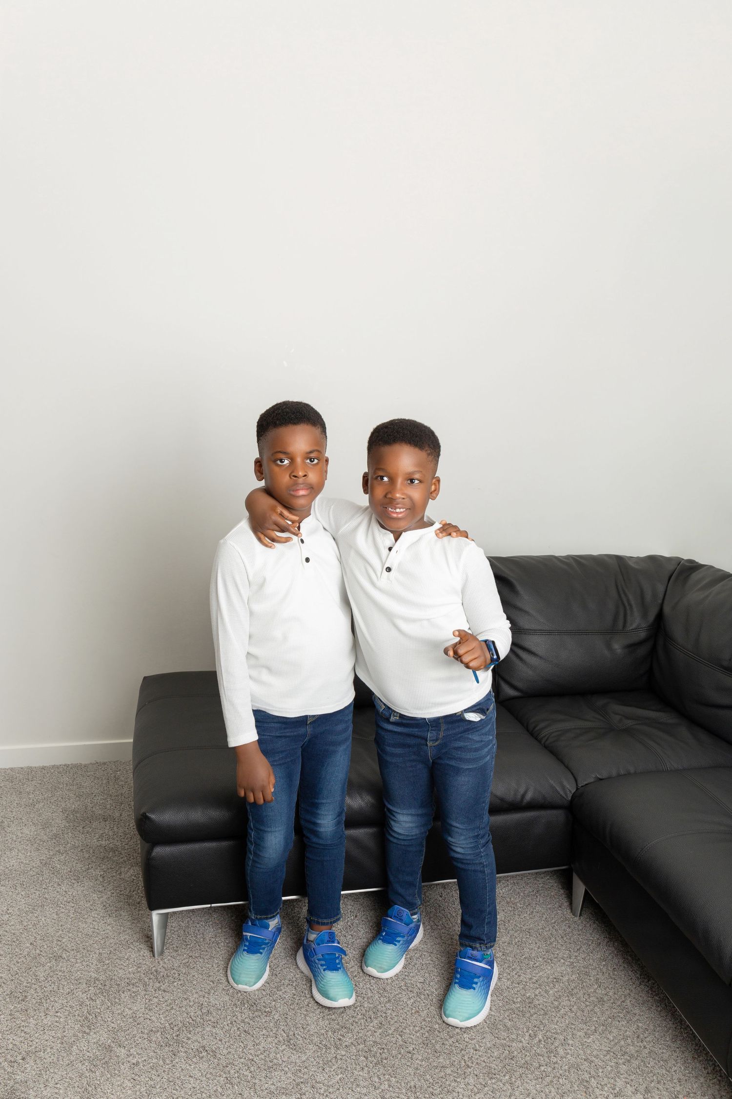 Two young twin boys in matching white shirts and jeans stand smiling by a black leather sofa.