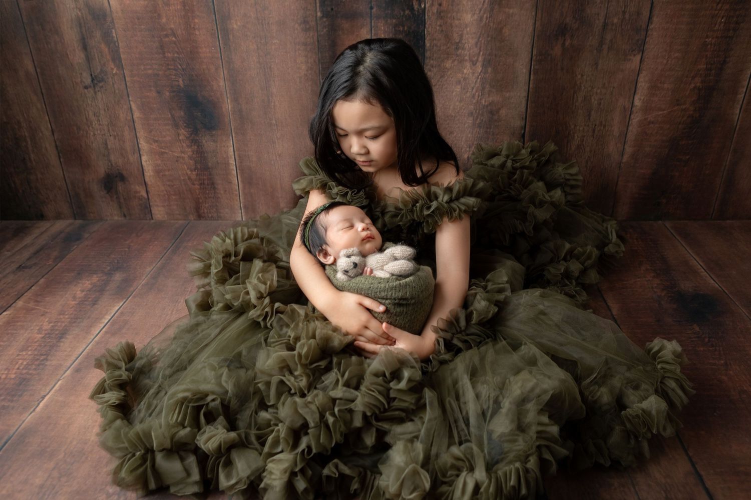 Young girl in olive tulle gown cradles swaddled newborn baby in professional studio portrait.