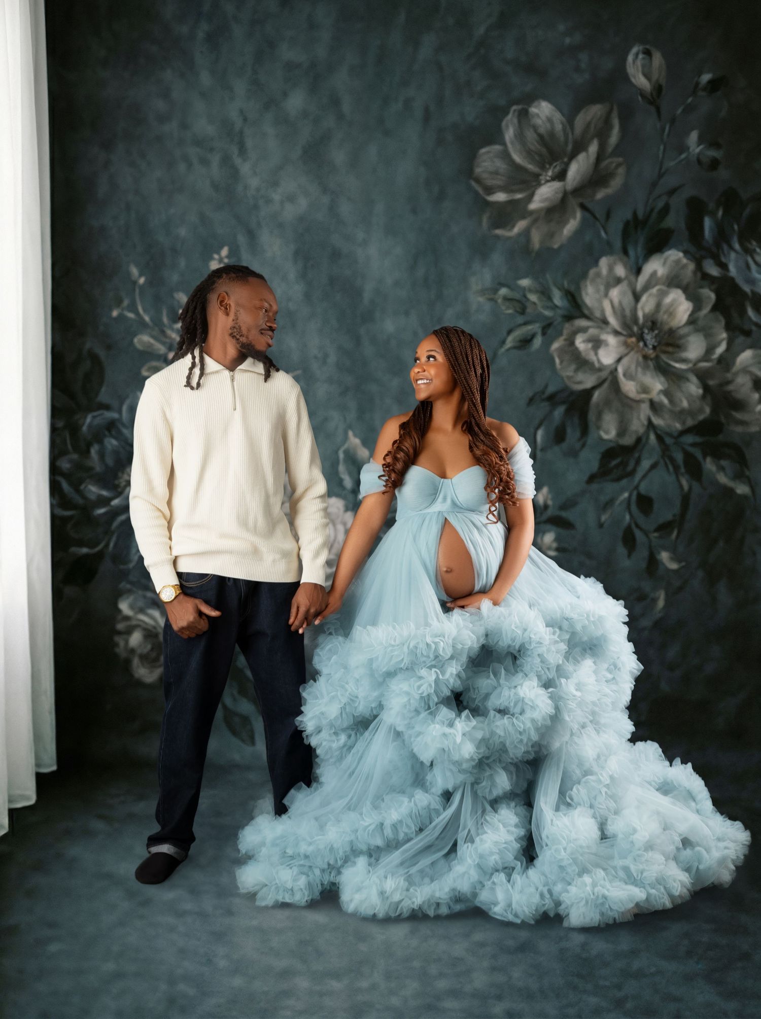 Couple posing for maternity photo, woman in blue tulle gown holding hands with man in cream sweater, floral backdrop.