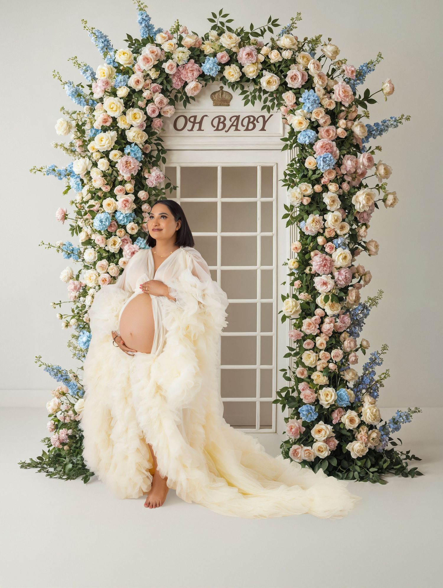 Pregnant woman in white ruffled gown poses under floral arch with pink and blue flowers and OH BABY sign.