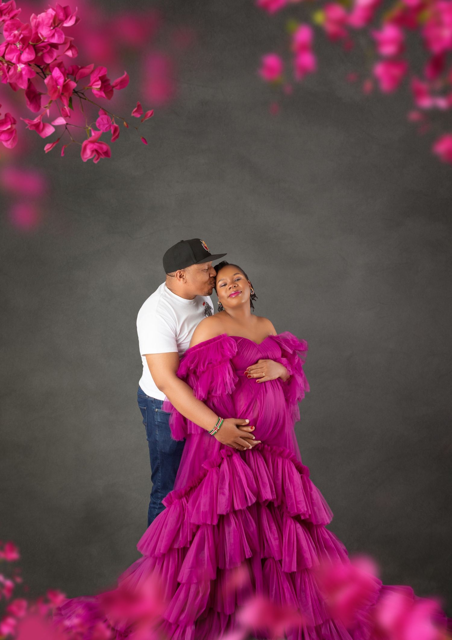 Couple in maternity photo shoot, woman in magenta ruffle gown, man kissing her cheek, pink flowers, gray backdrop.