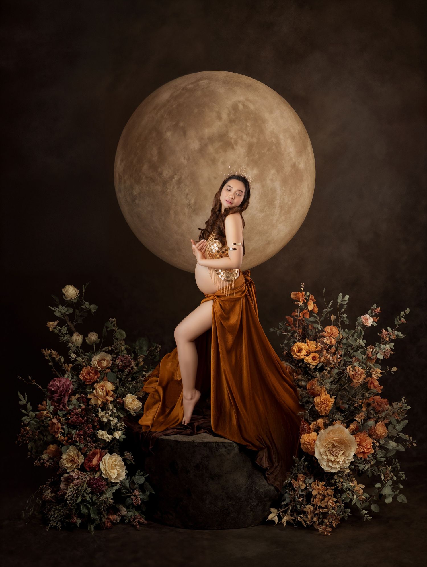 Pregnant woman in rust satin skirt posed before a large moon backdrop surrounded by florals.