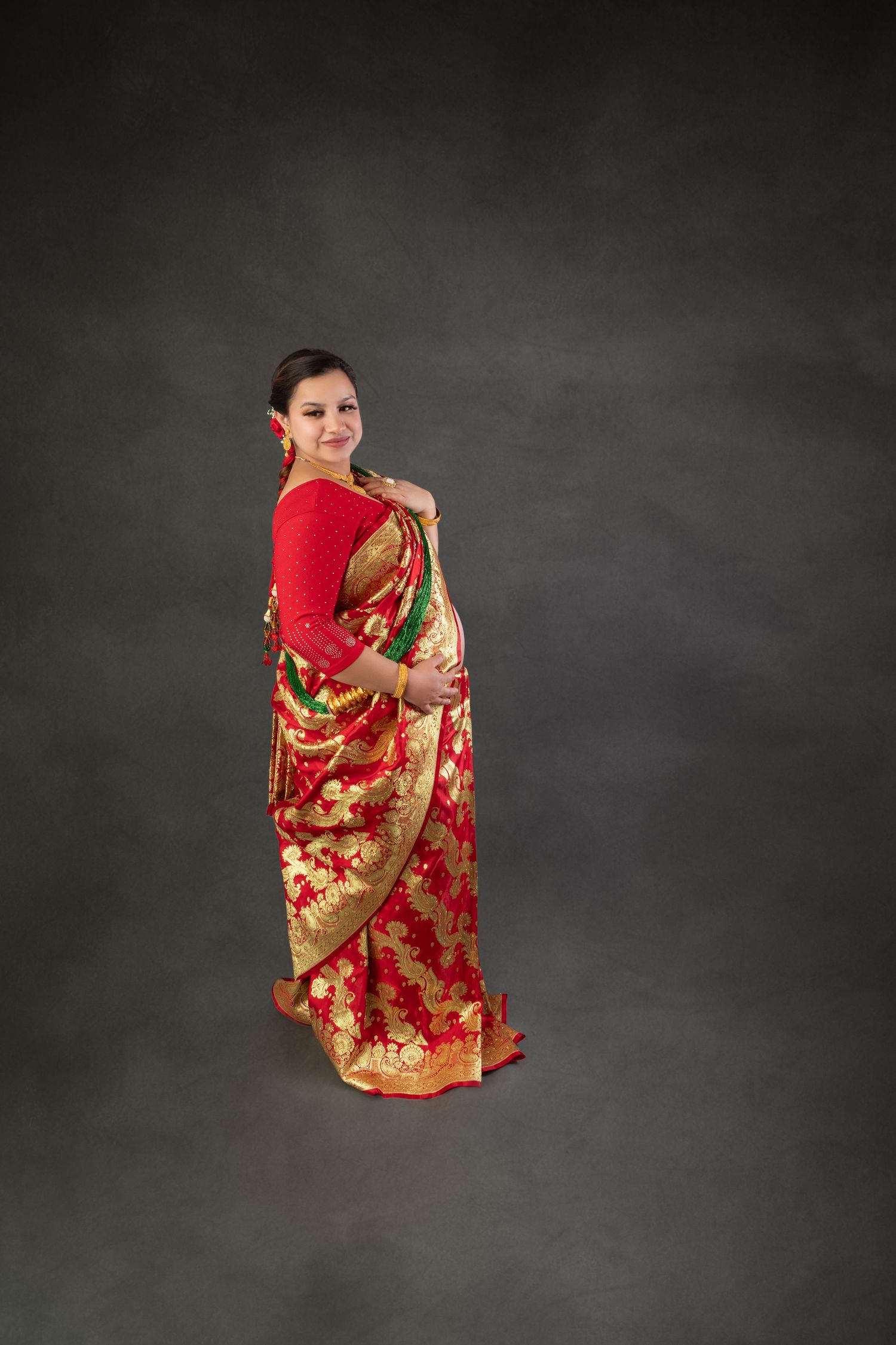 Pregnant woman posing in elegant red and gold traditional Indian saree during maternity photoshoot.