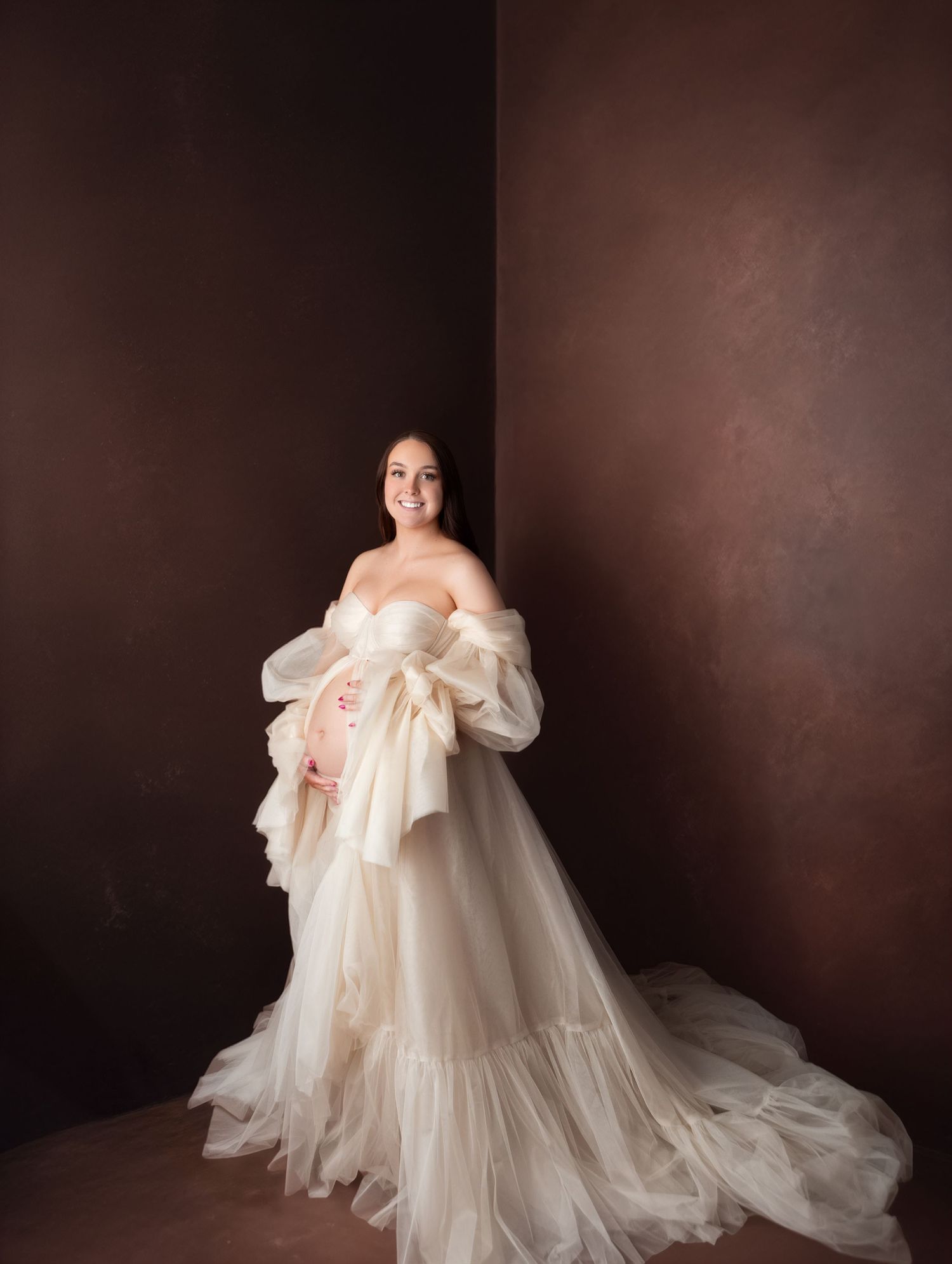 Expecting mother in off-shoulder cream tulle maternity gown poses against warm brown studio backdrop.