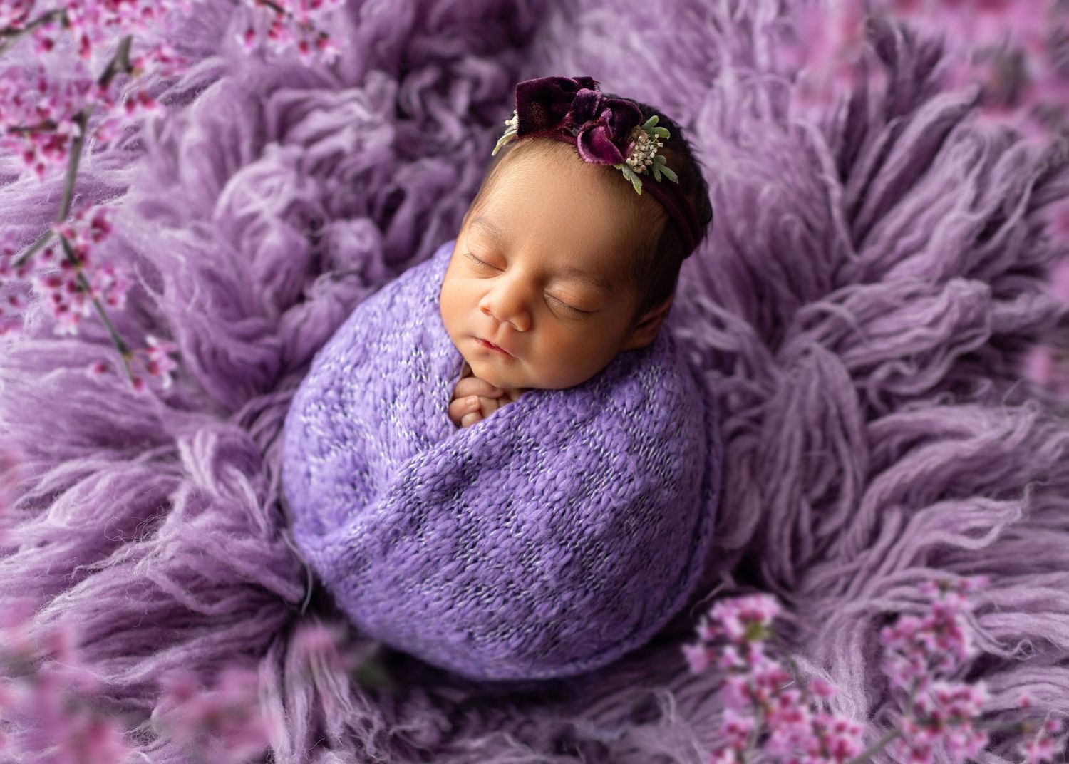 Newborn baby wrapped in purple knit blanket with floral headband on lavender fur backdrop.