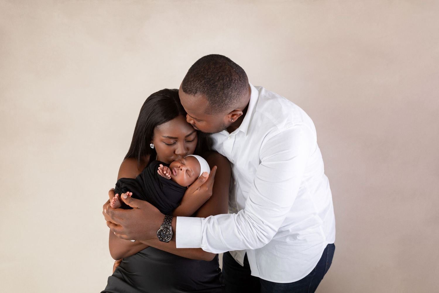 Loving parents lean in to kiss their swaddled newborn baby against a warm neutral backdrop.