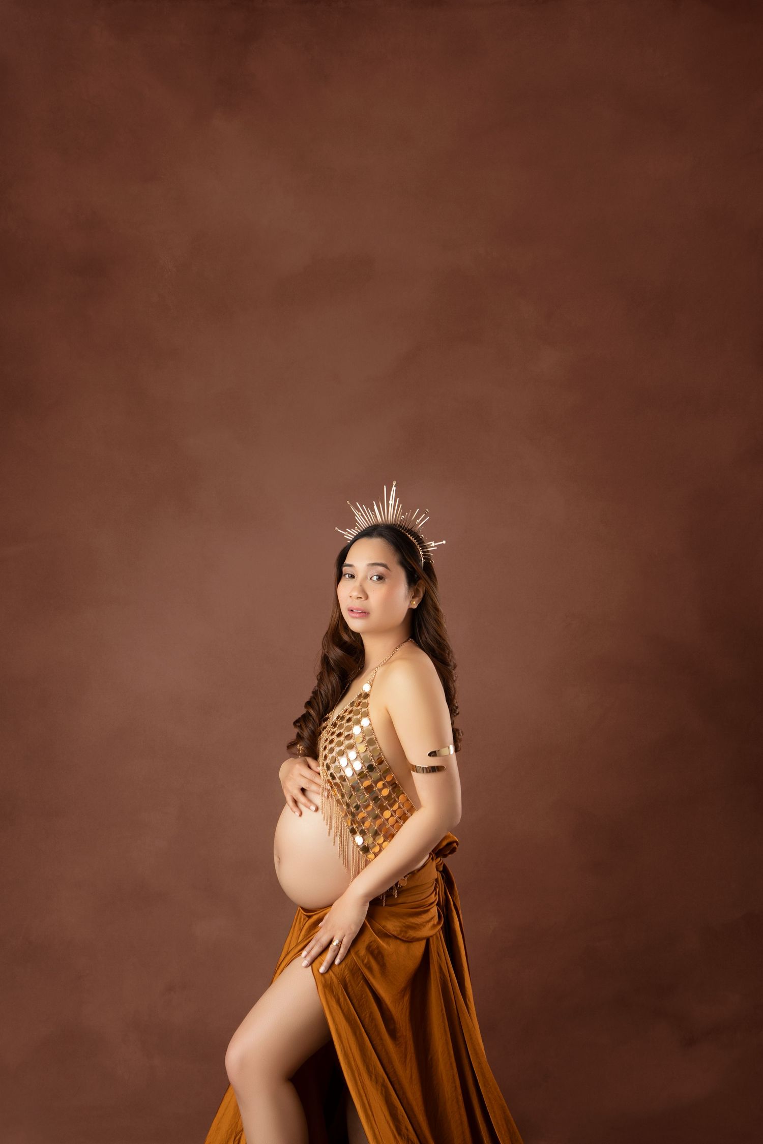 Pregnant woman wearing gold sequin top and burnt orange velvet skirt with crystal crown in studio portrait.