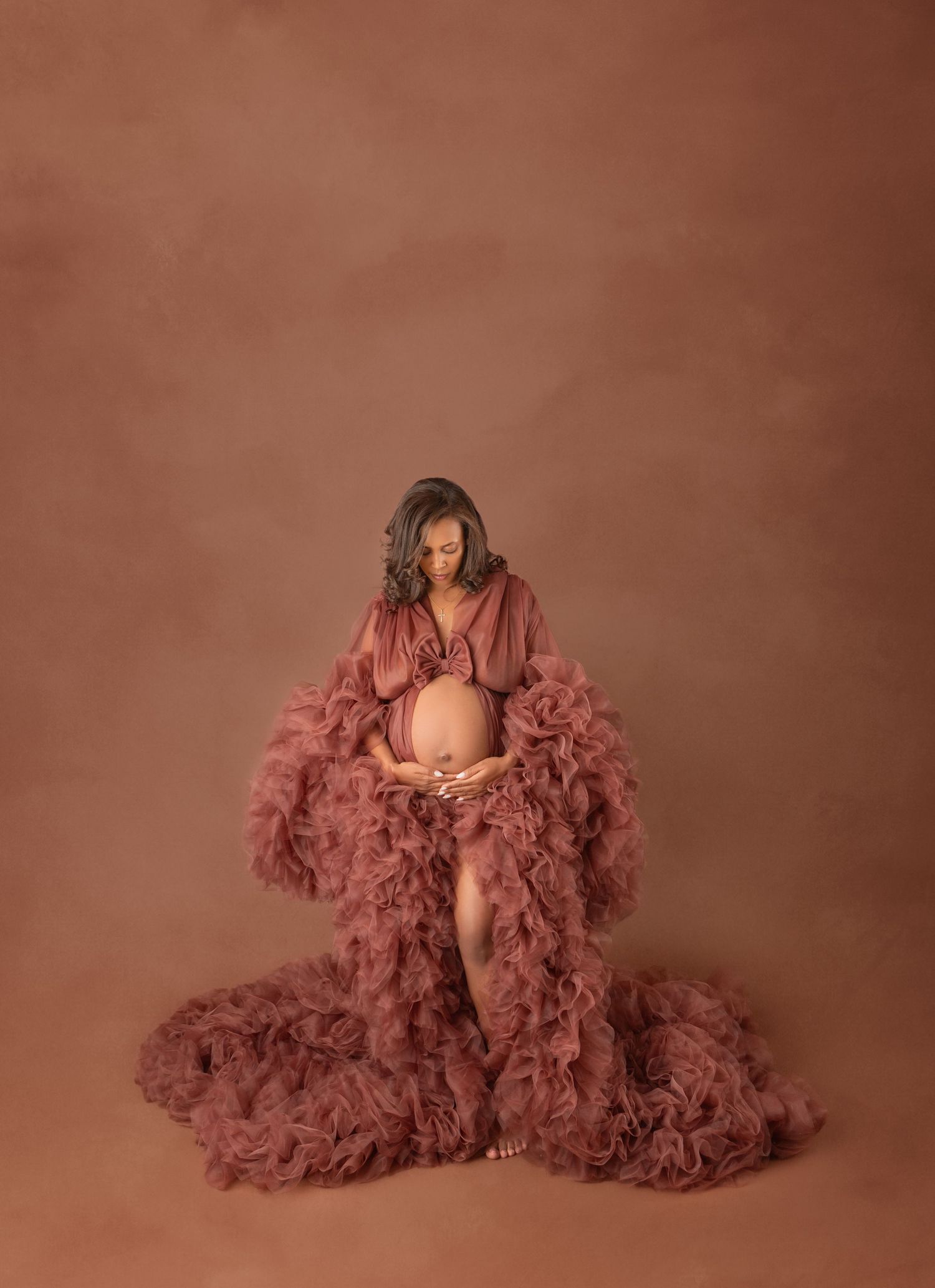Pregnant woman in mauve ruffled tulle gown looks down tenderly at her belly in a studio maternity portrait.