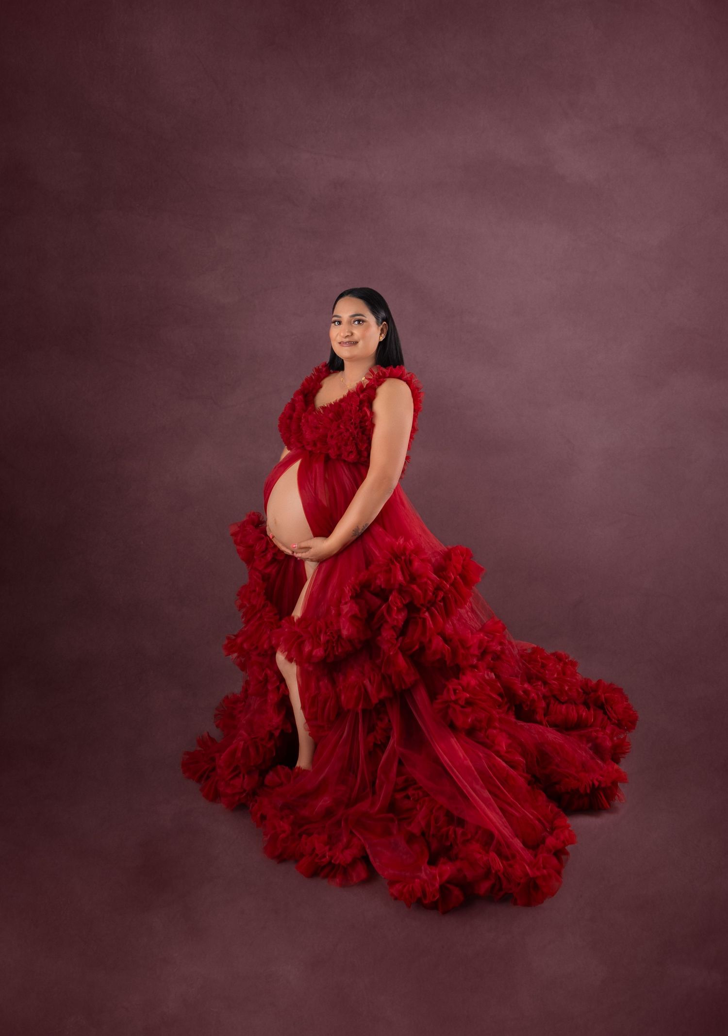 Pregnant woman in elegant red ruffled tulle maternity gown posing gracefully against a mauve studio backdrop.