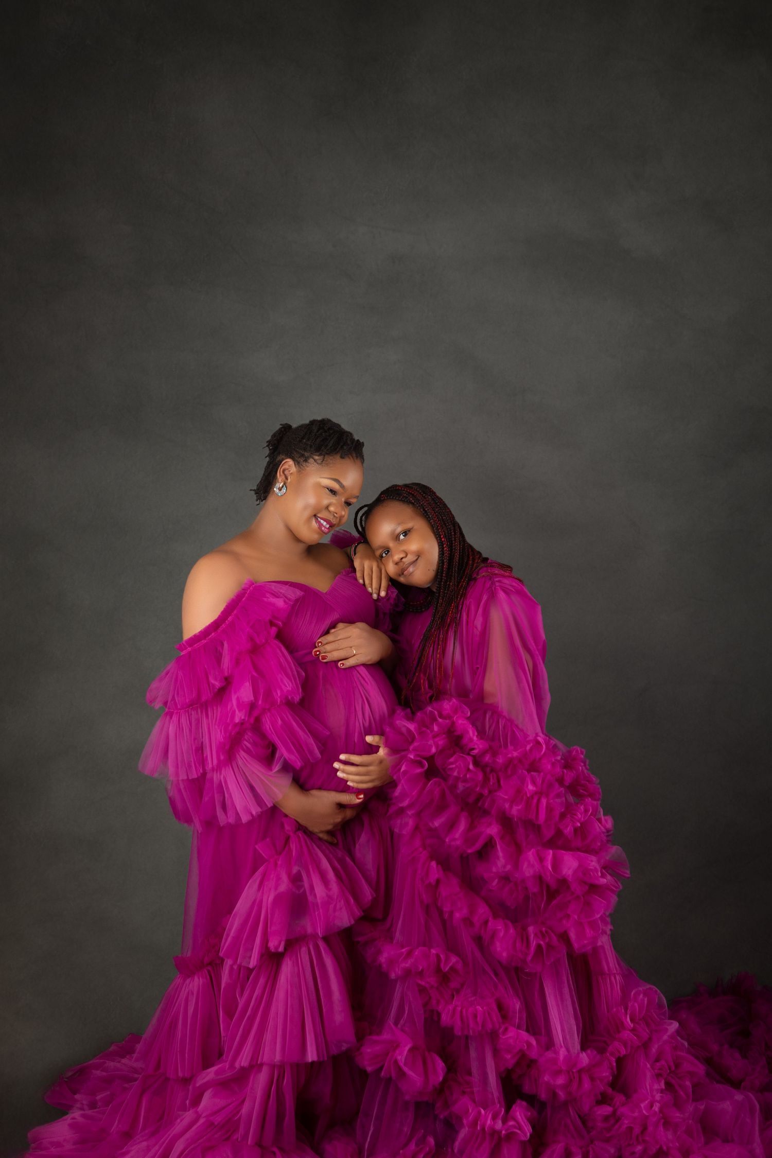 Two women in matching magenta ruffle gowns pose together in a maternity portrait against a gray studio backdrop.