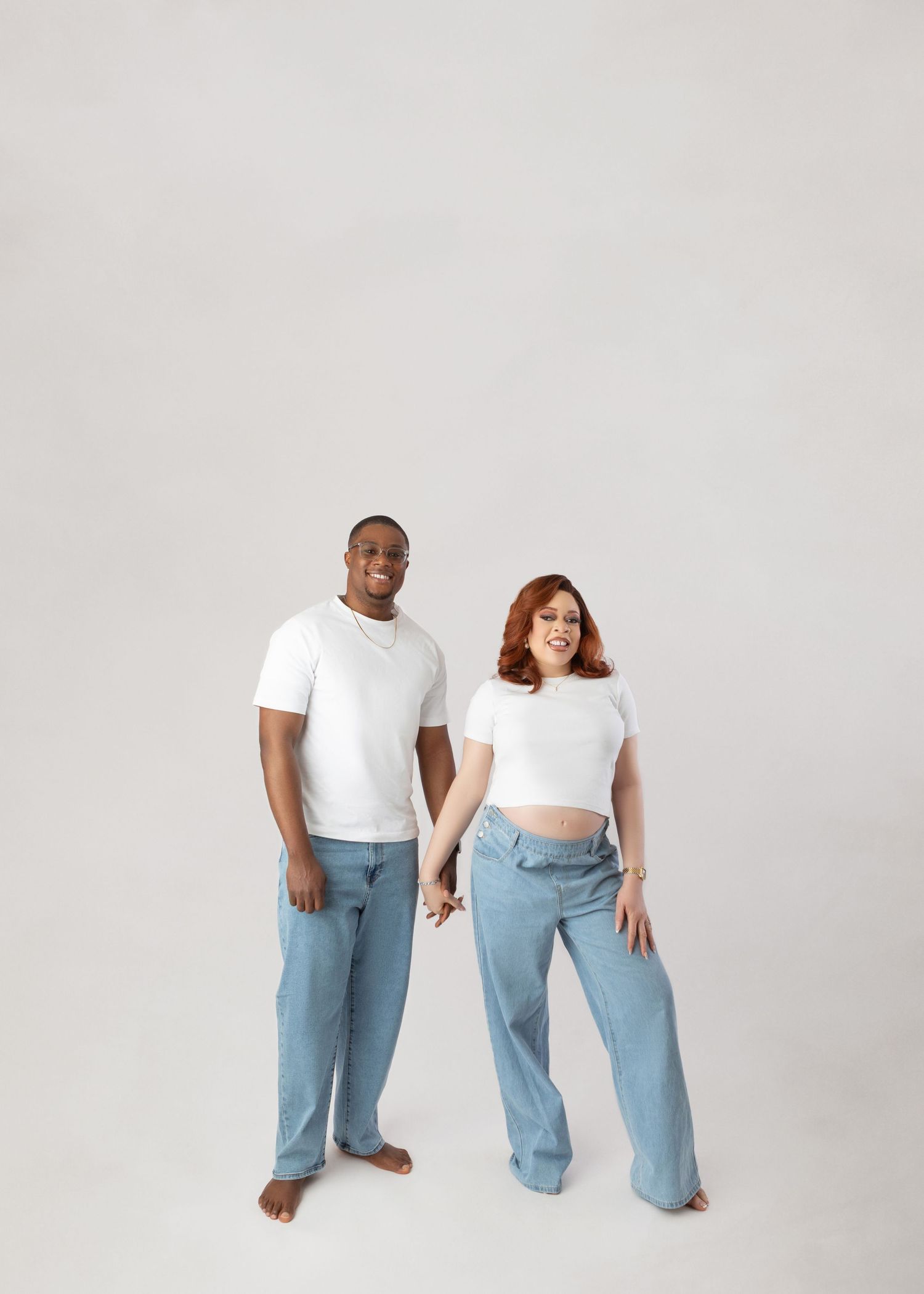 Smiling expecting couple in matching white tees and blue jeans holding hands against a light gray background.
