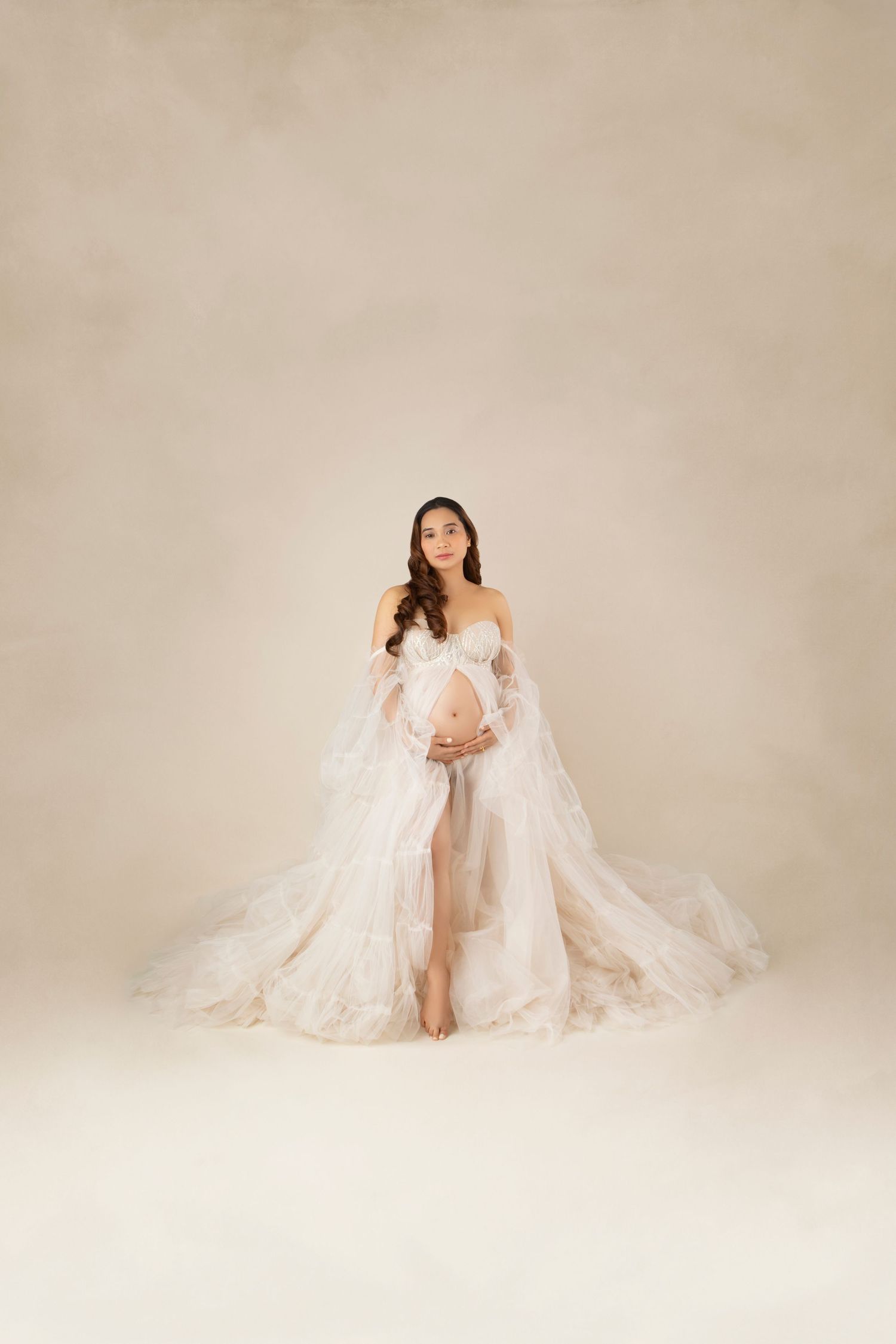 A pregnant woman in a flowing white floral tulle gown stands elegantly against a neutral background.