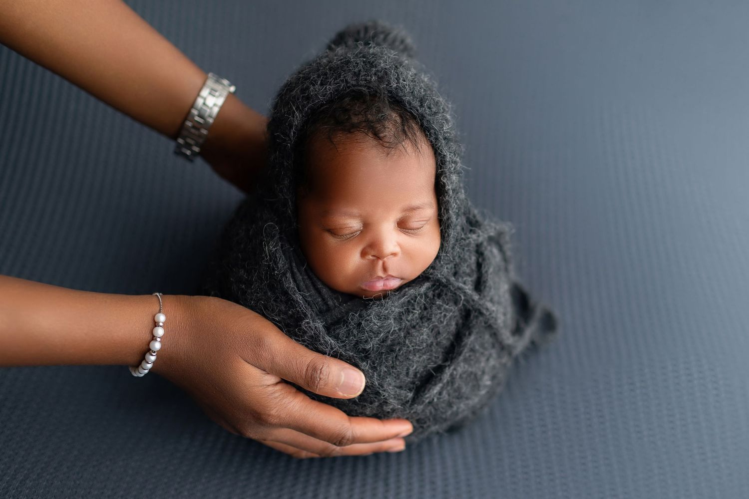 Sleeping newborn wrapped in gray knit fabric cradled gently by adult hands on gray backdrop.