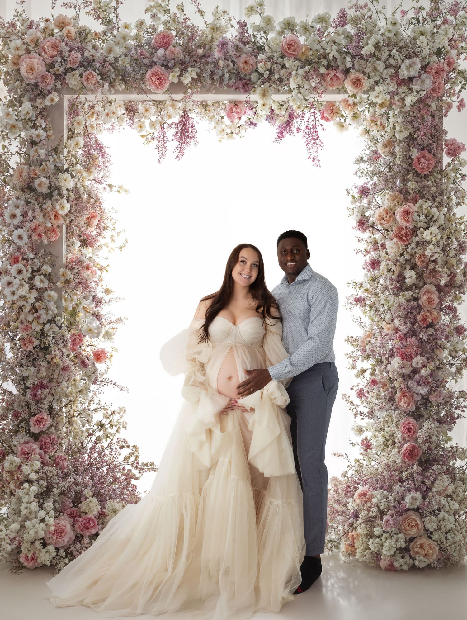 Couple maternity portrait under floral arch, woman in cream ruffled tulle gown.