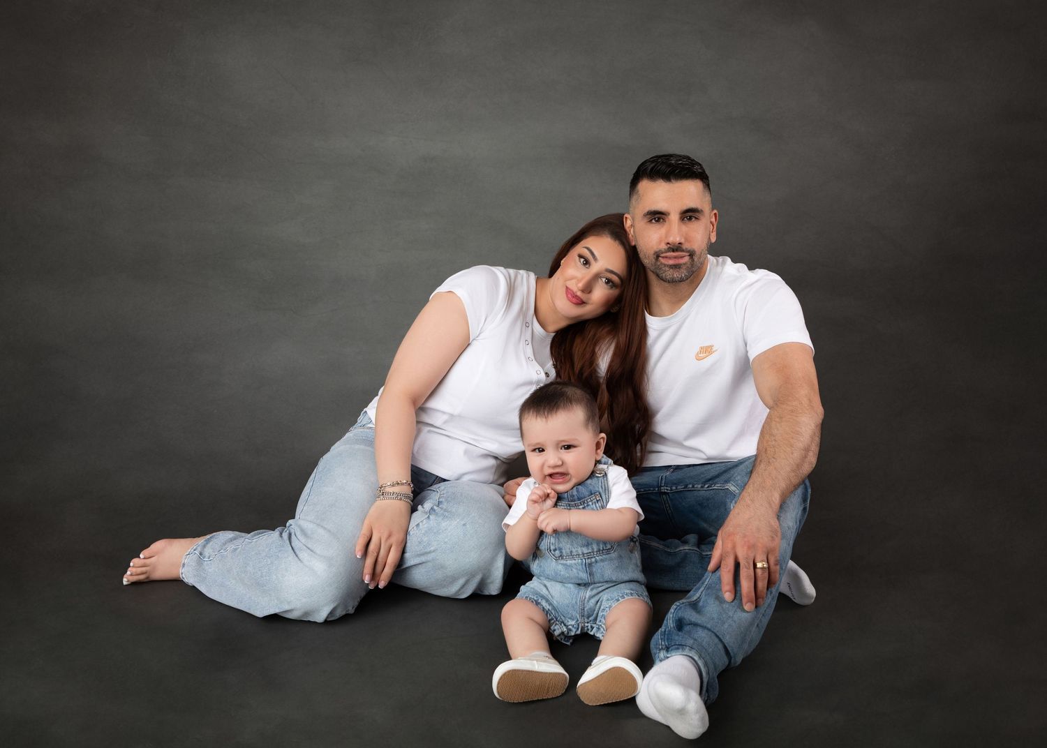 Family sitting casually with baby in denim overalls against dark grey backdrop.