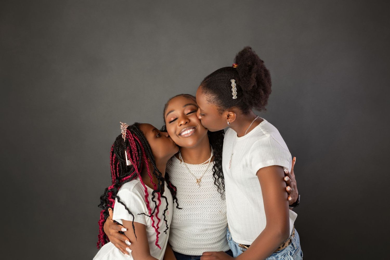 Three Black sisters sharing sweet kisses on cheeks in white tops against dark gray background.