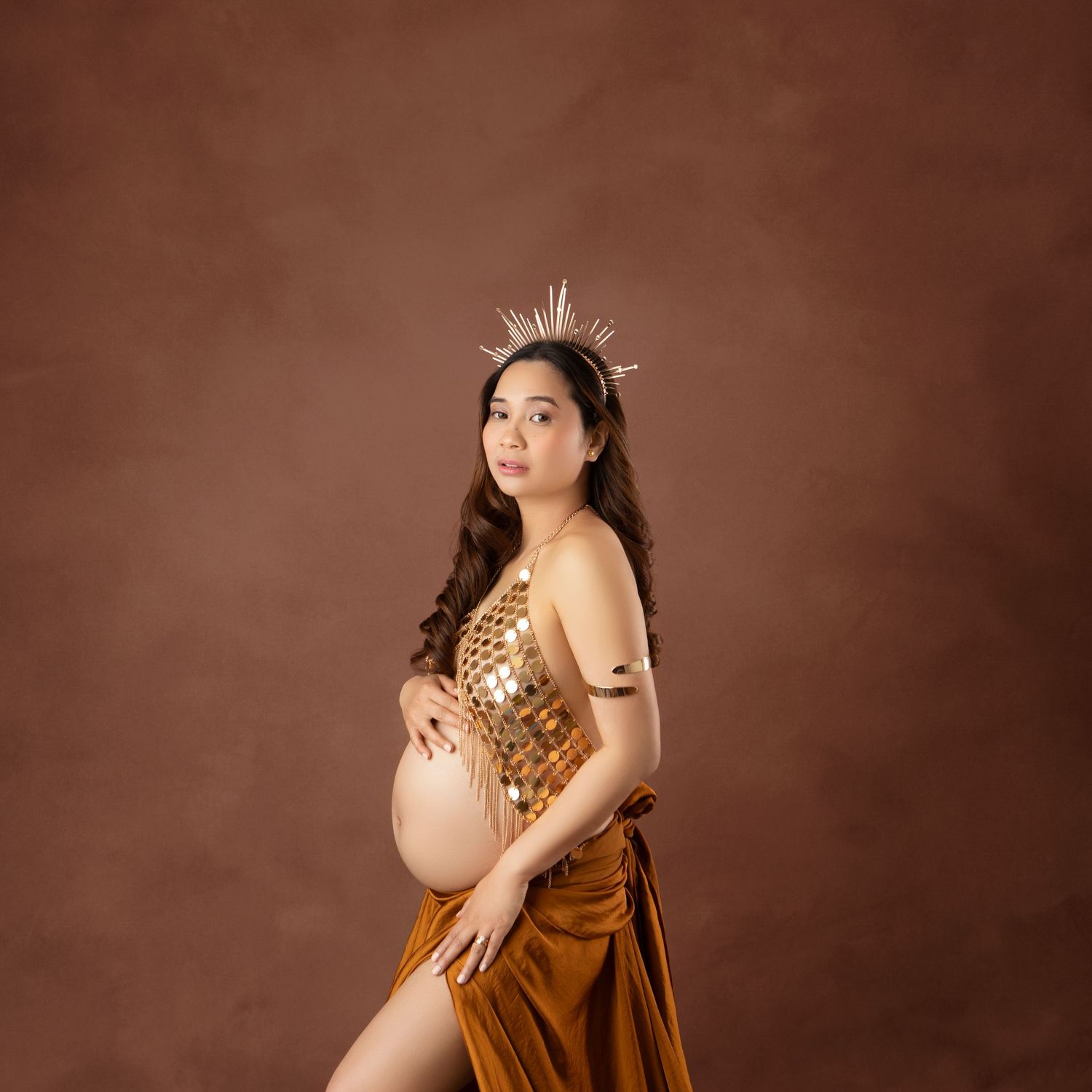 Pregnant woman in golden sequin top and rust velvet skirt wearing crown poses for maternity photo session.