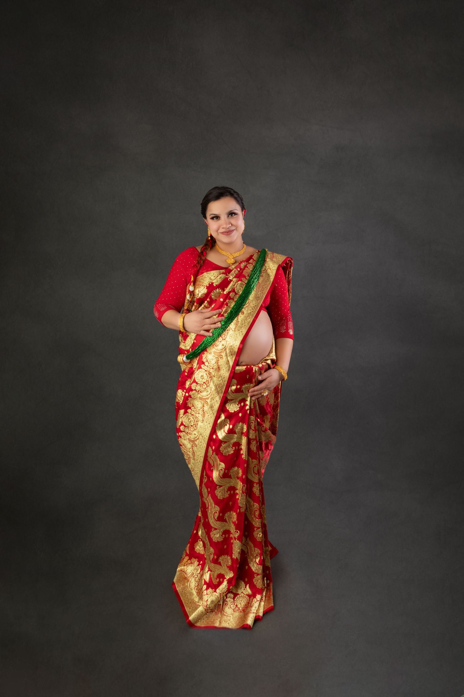 Pregnant woman in red and gold traditional Indian saree posing gracefully against a dark gray background.