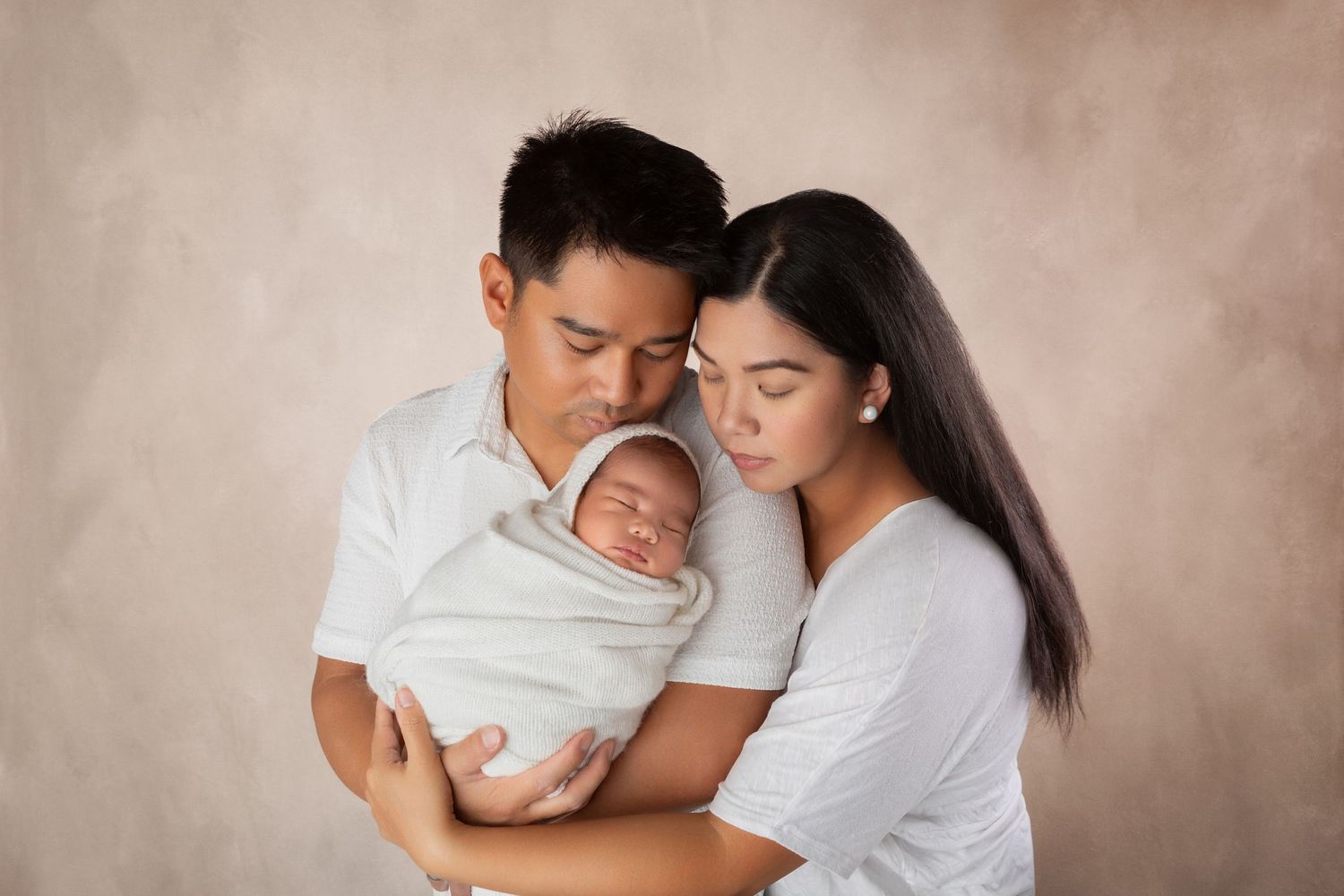 A couple in white outfits tenderly holds their sleeping newborn baby wrapped in white against a neutral backdrop.