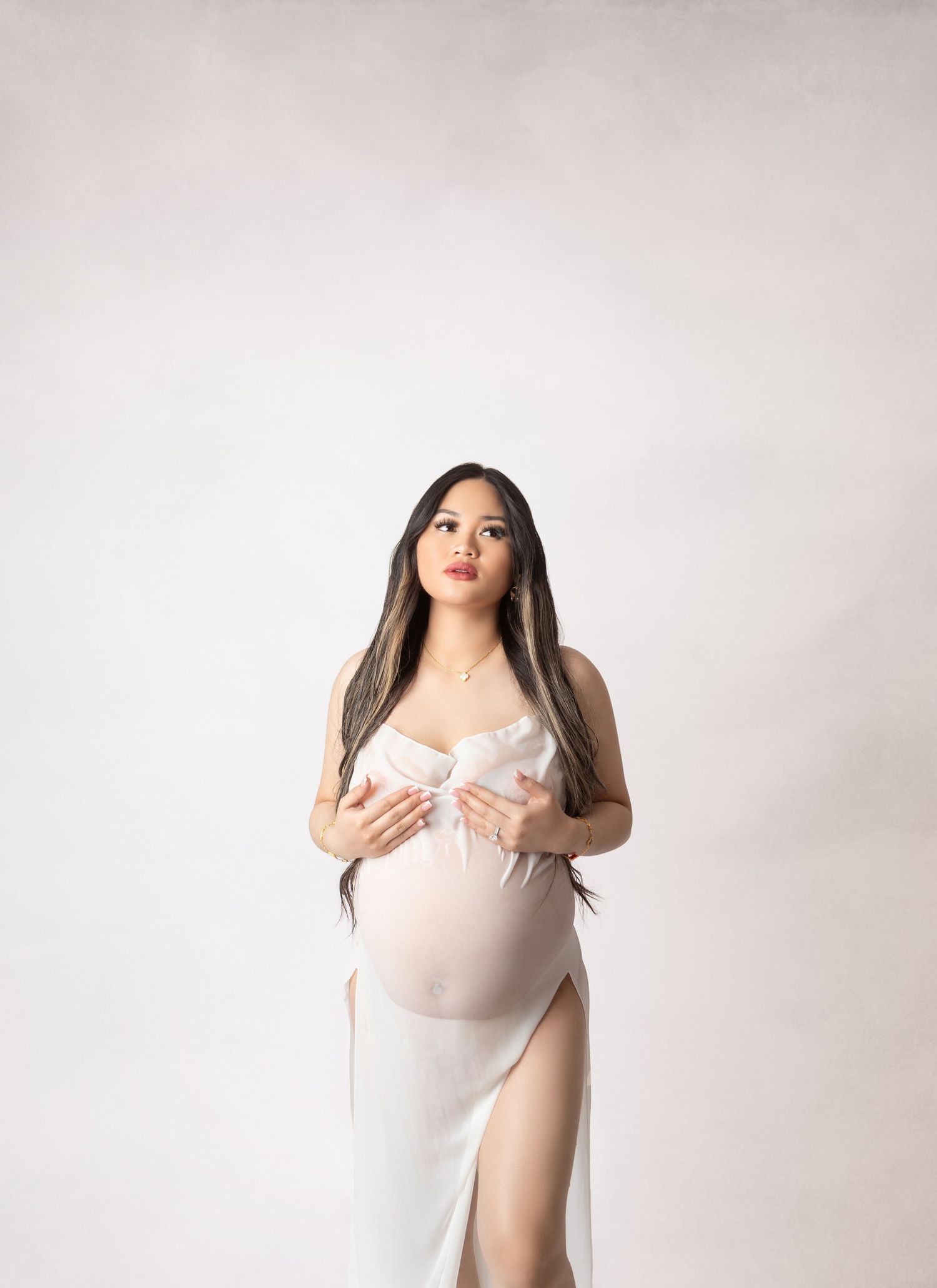 Pregnant woman in white satin maternity gown cradling baby bump, long dark hair, studio portrait.