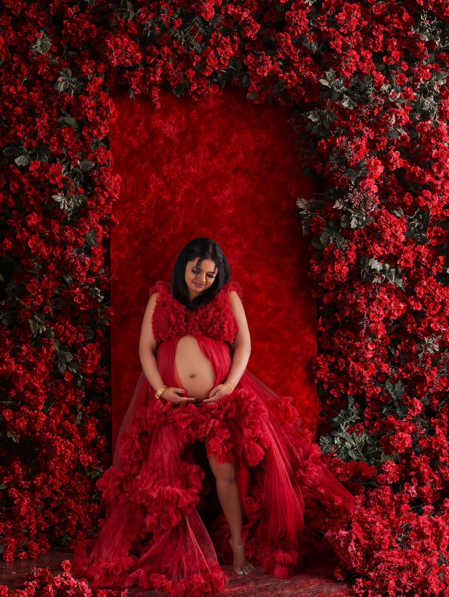Pregnant woman in red tulle gown poses in floral arch surrounded by lush red roses for maternity photo shoot.