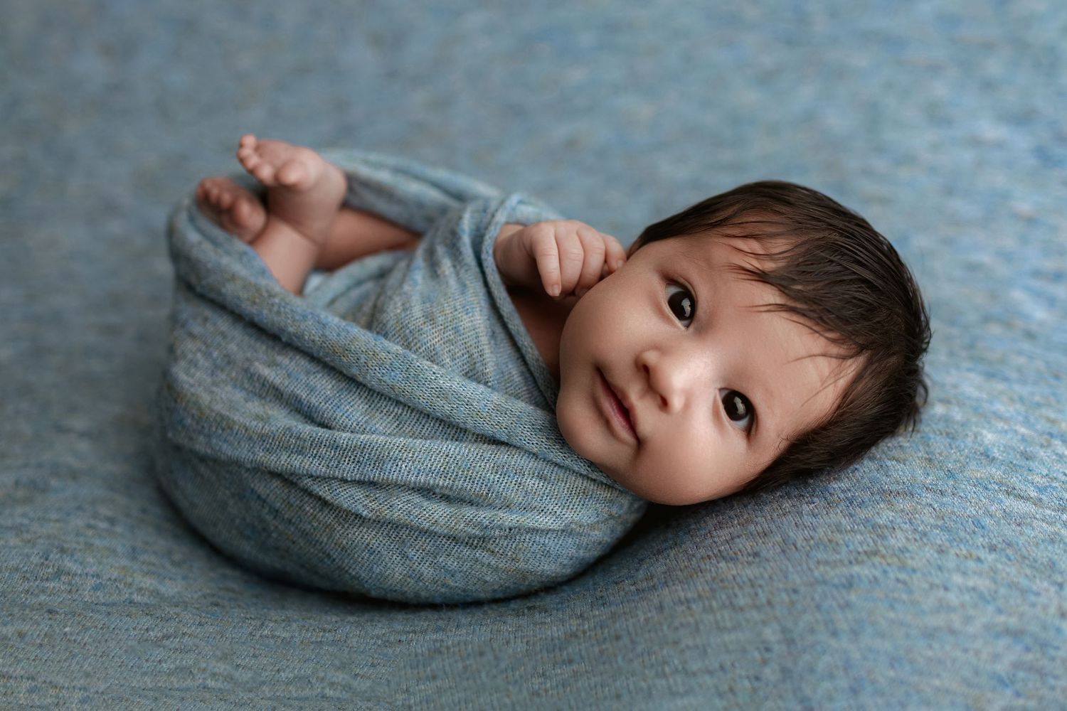 Awake newborn baby swaddled in a soft gray wrap lying on a blue blanket during a newborn photo session.