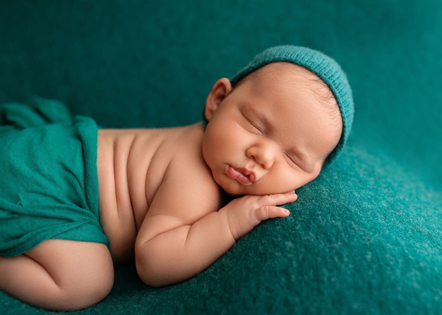 Peaceful newborn baby sleeping on teal blanket wearing knit hat, professional newborn photography portrait.