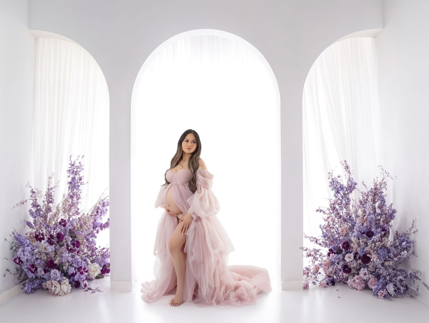 Pregnant woman in pink tulle gown posing under white arches surrounded by purple floral arrangements.