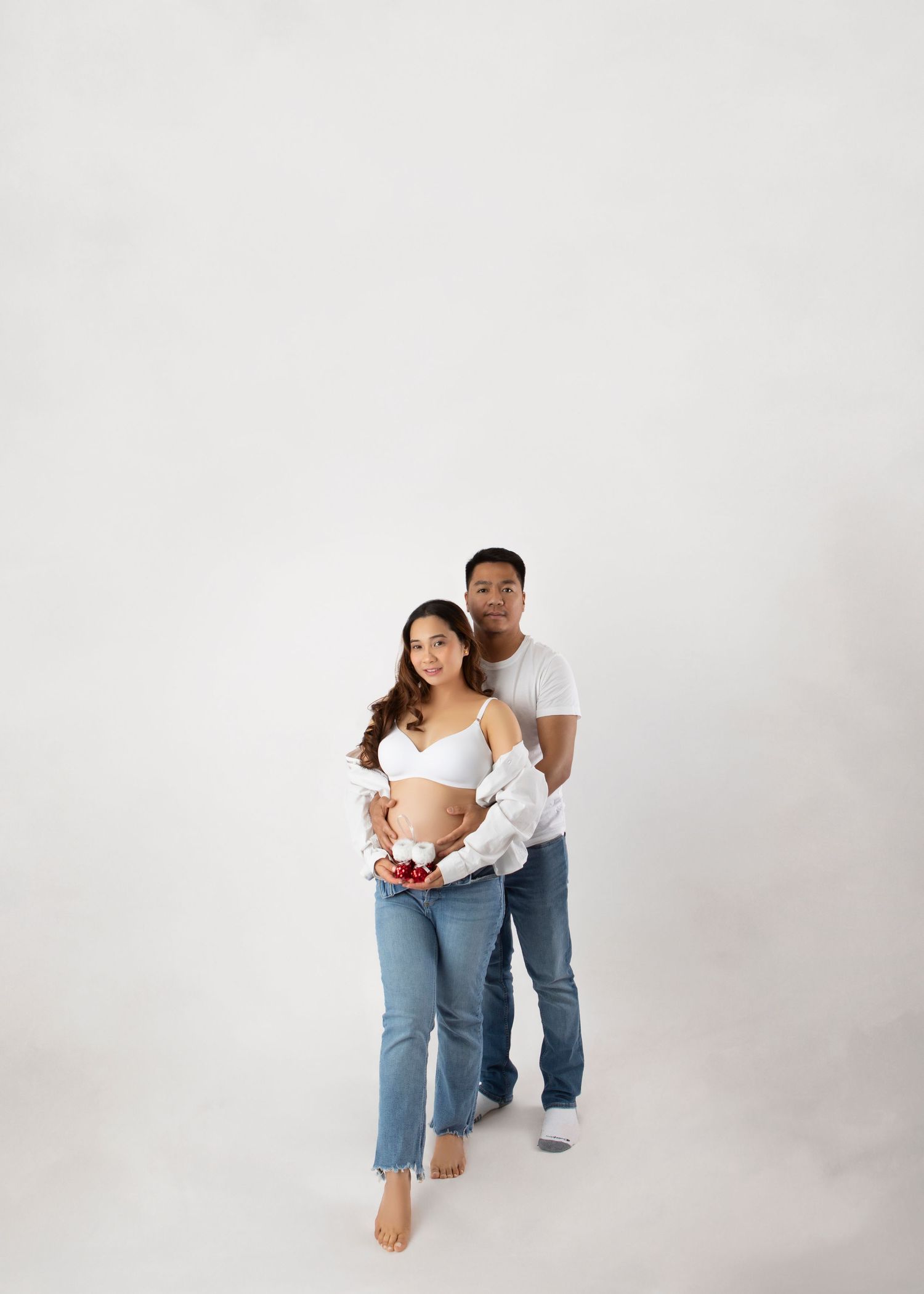 Pregnant couple in white tops and blue jeans pose together holding small red baby shoes on white backdrop.