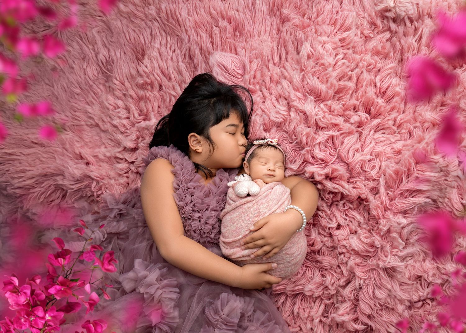 Big sister in purple dress kisses swaddled newborn sibling on pink fur surrounded by flowers.