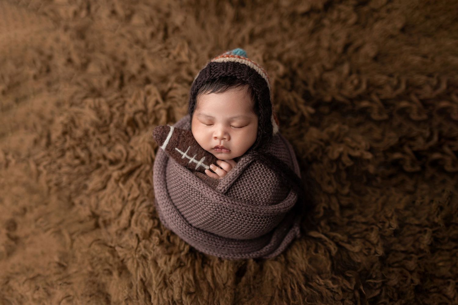 Newborn baby wrapped in brown knit swaddle wearing a colorful bonnet on fluffy brown rug.