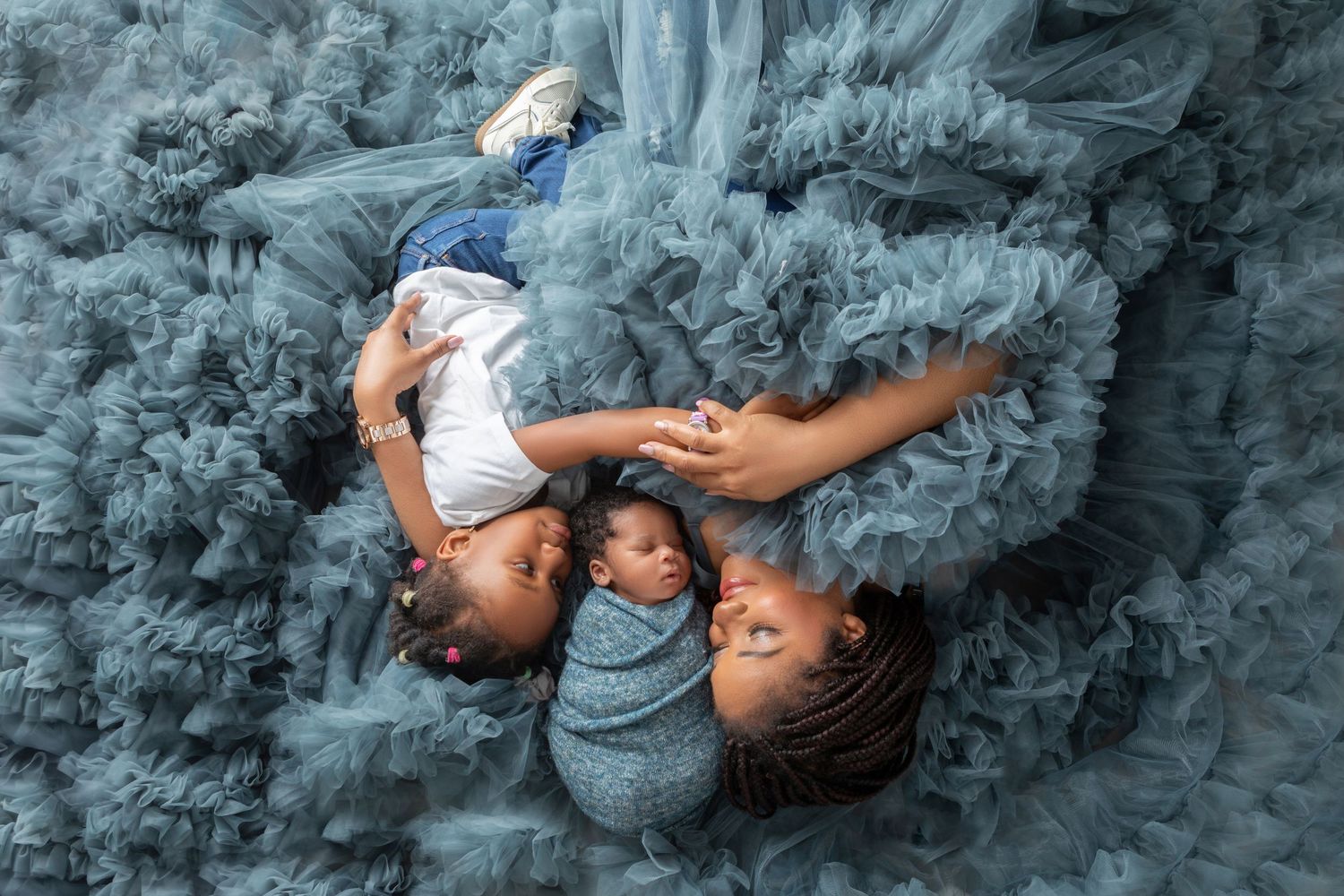 Mother in blue tulle gown lies with newborn baby and toddler on matching ruffled fabric in aerial portrait.