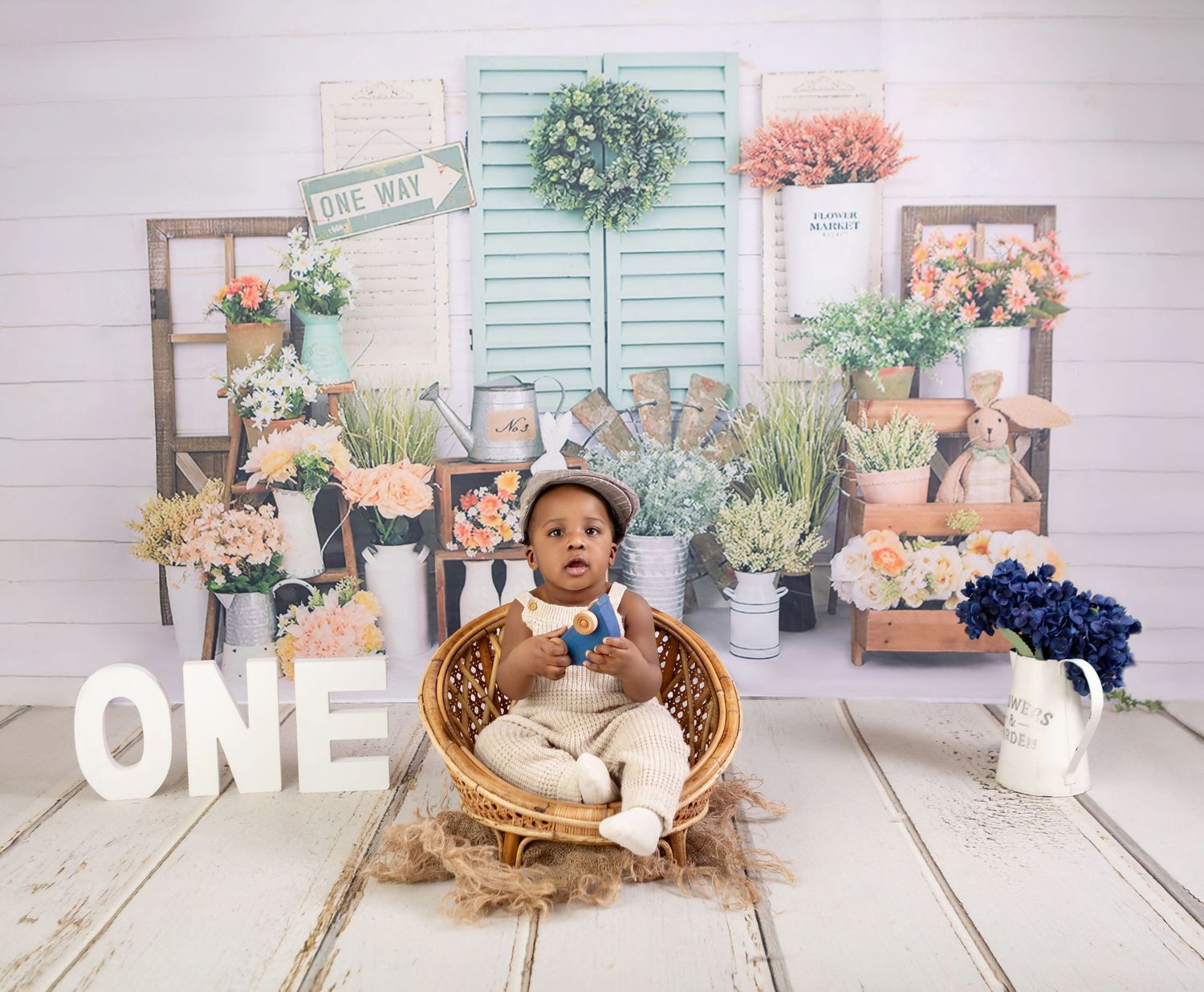 Baby's first birthday photo shoot with floral garden backdrop, sitting in wicker chair surrounded by plants and ONE letters.