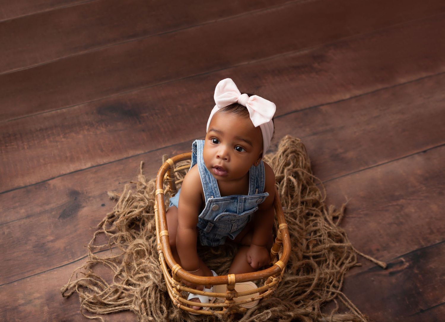 Adorable baby girl sitting in wicker basket wearing denim romper and pink bow headband on wood floor.