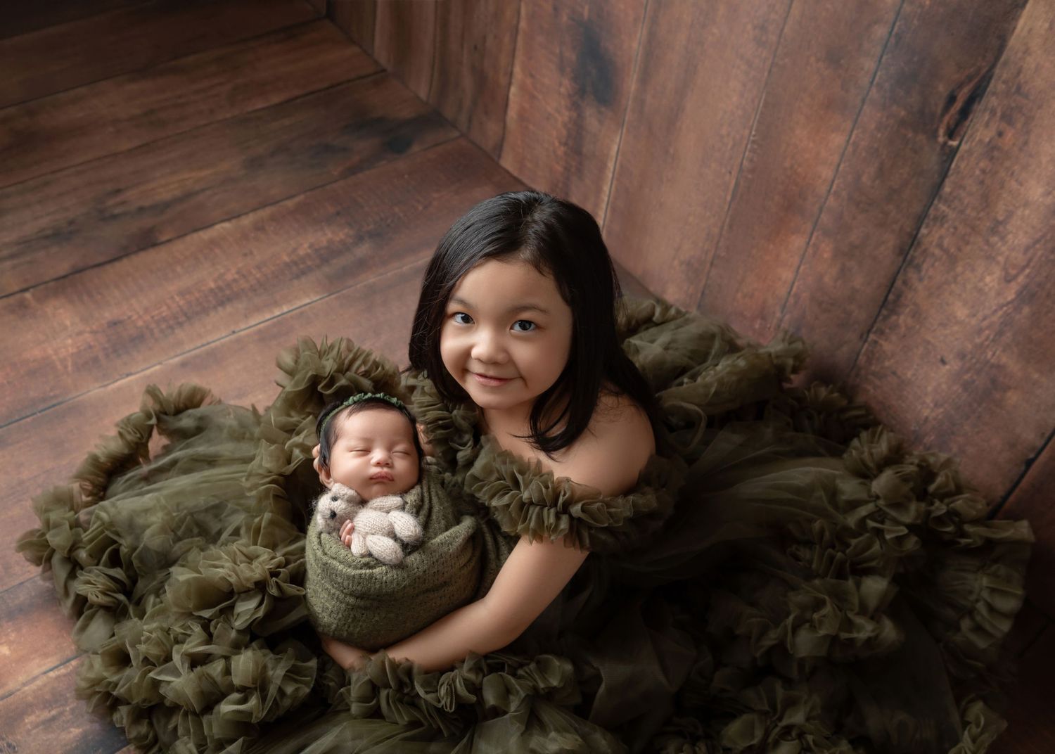 Young girl in olive tulle dress holds swaddled newborn sibling on rustic wood floor.
