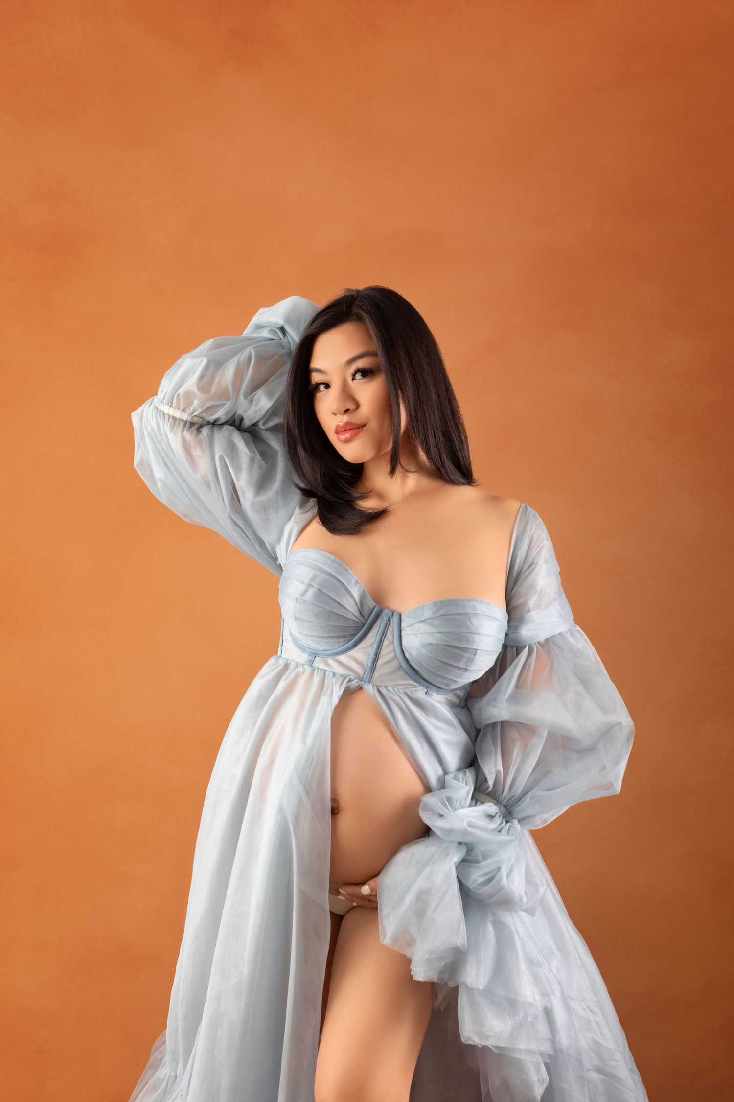 Pregnant woman in blue tulle robe and corset posing against orange background.