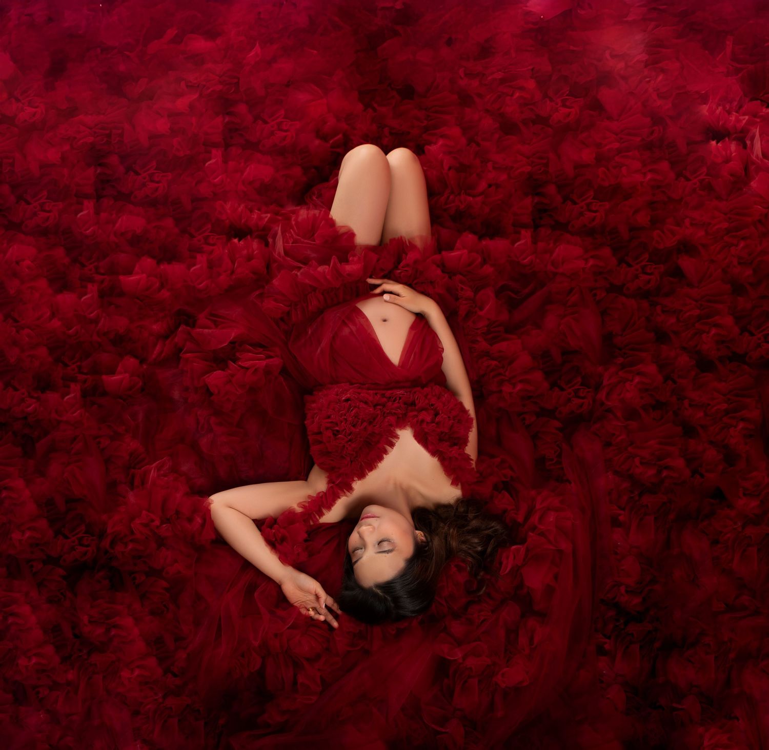 Pregnant woman lying on a lush bed of deep red flowers wearing a red ruffled maternity gown.