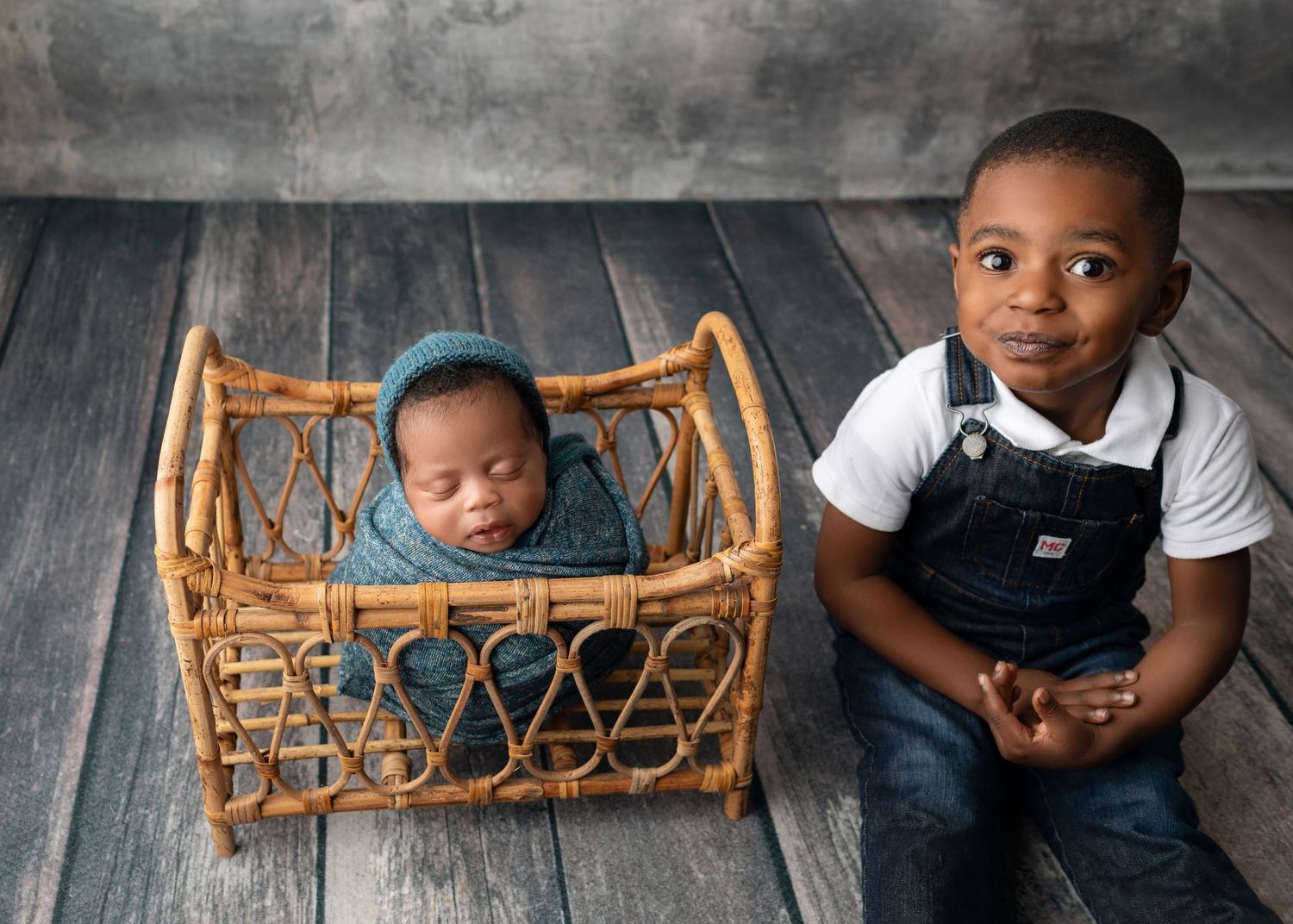 Newborn baby sleeping in wicker bassinet beside proud toddler brother in denim overalls on rustic wood floor.