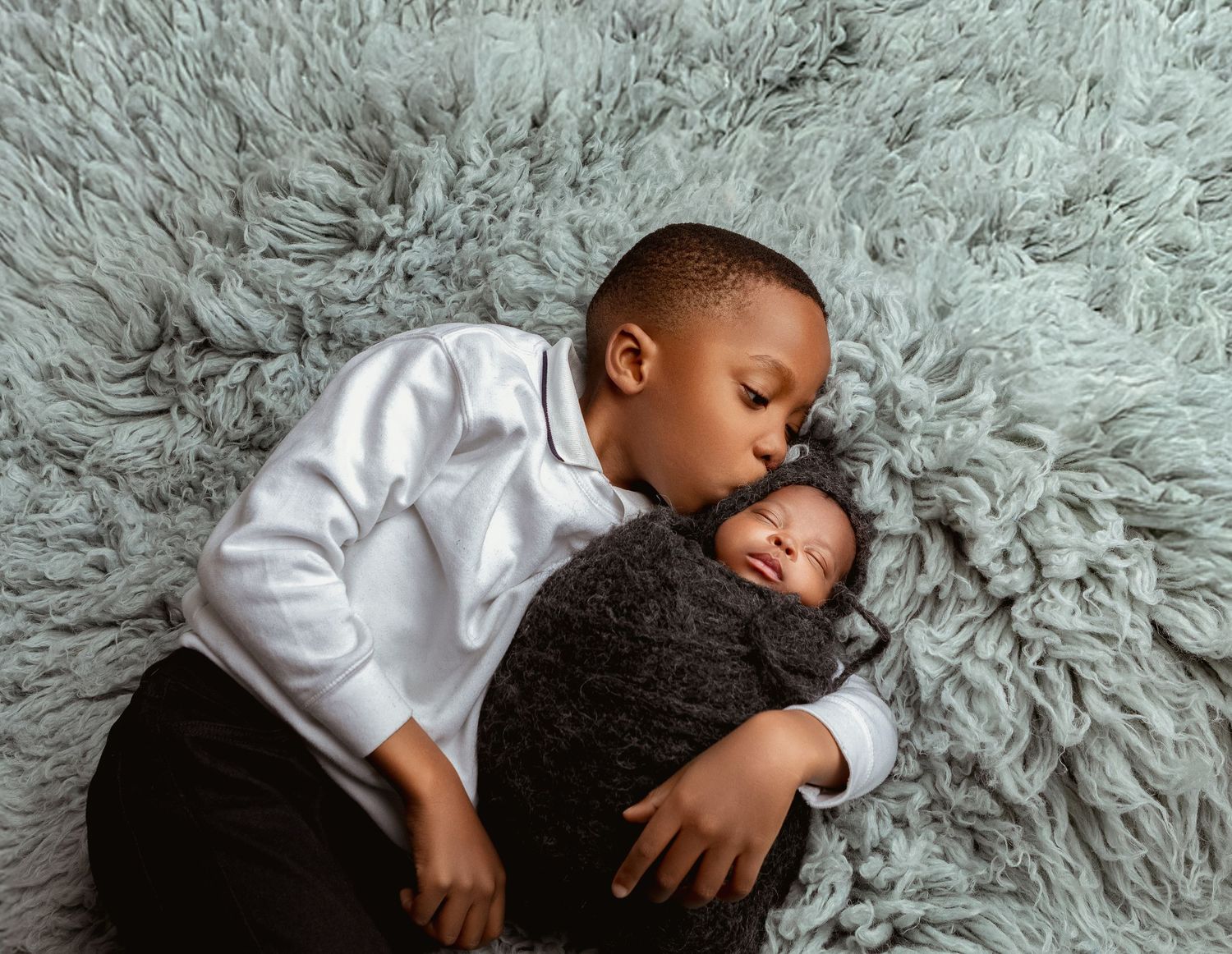 A young boy in a white shirt tenderly cuddles his sleeping newborn sibling on a fluffy gray rug.