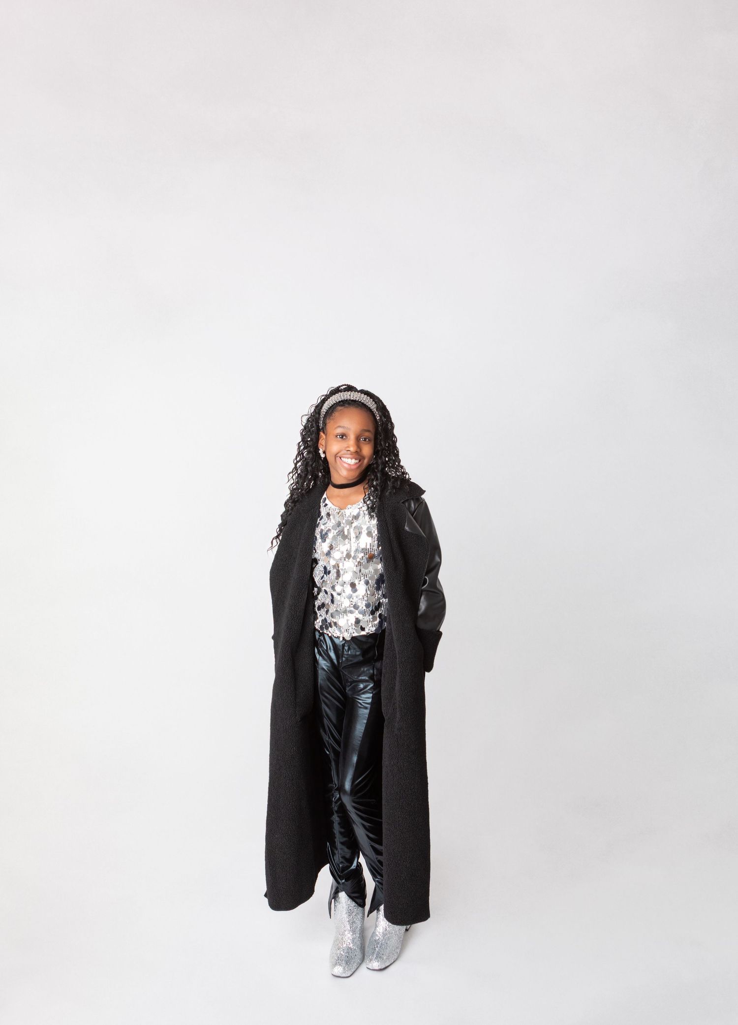 Young girl in long black coat, floral top, metallic pants, and silver boots smiling against white background.