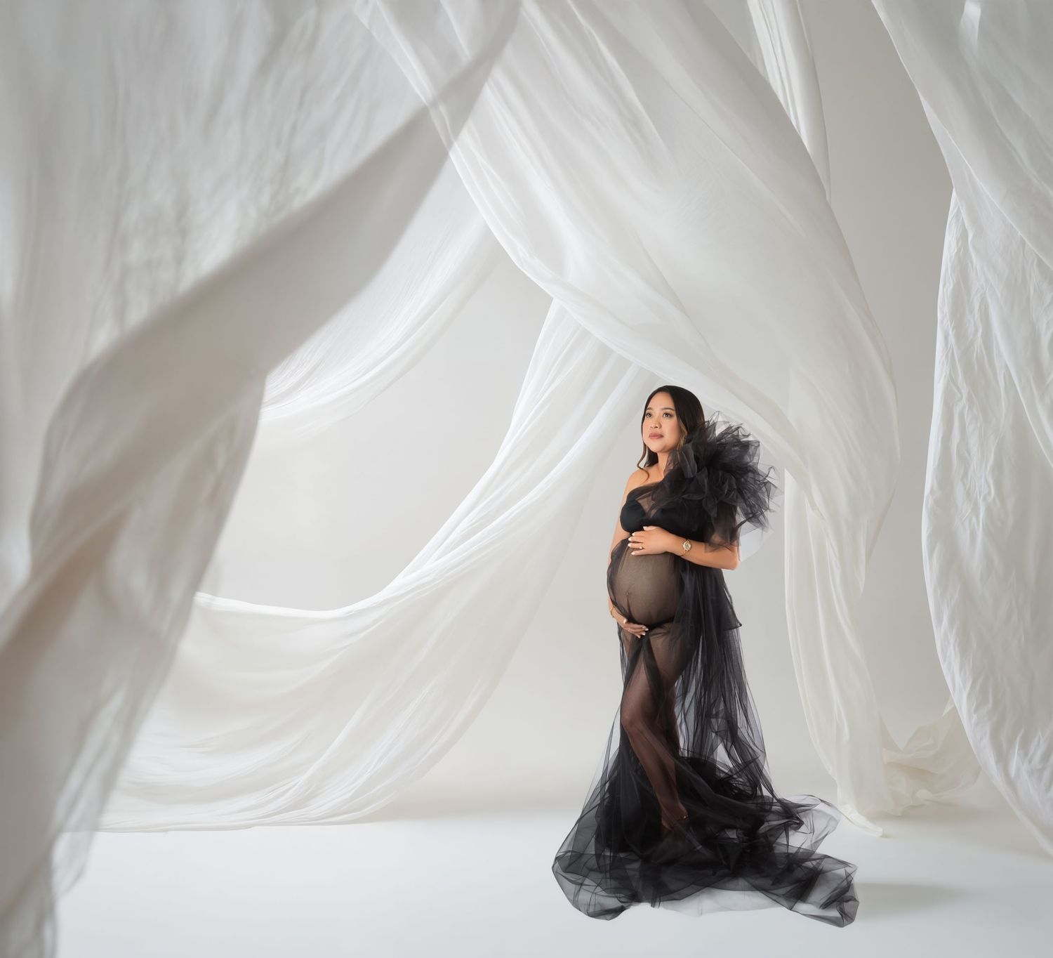 Pregnant woman in black sheer maternity gown poses gracefully among flowing white drapes in a bright studio setting.