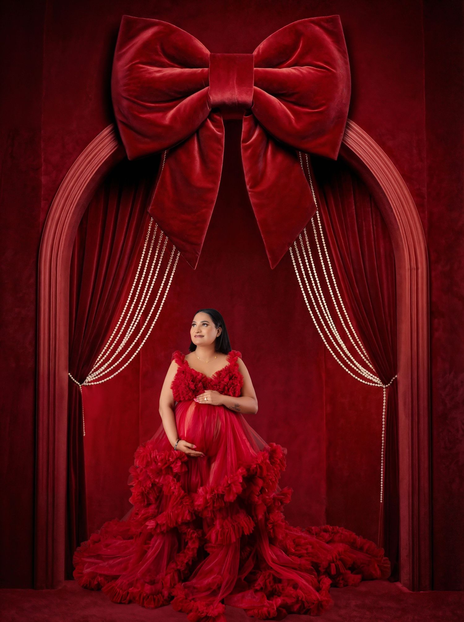 Pregnant woman in red ruffled maternity gown poses under ornate red velvet arch with large decorative bow.