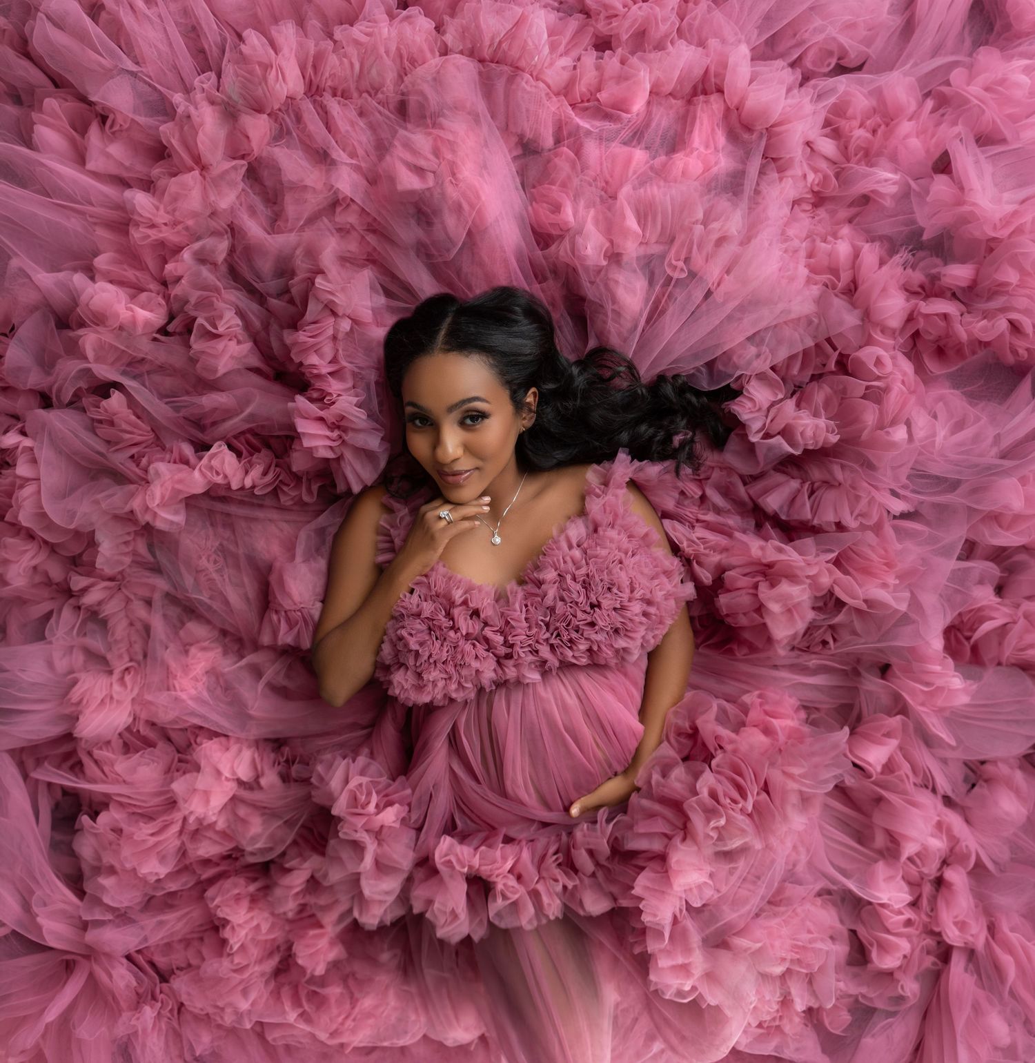 Pregnant woman in voluminous pink tulle maternity gown lying amid ruffled fabric, elegant maternity photography.